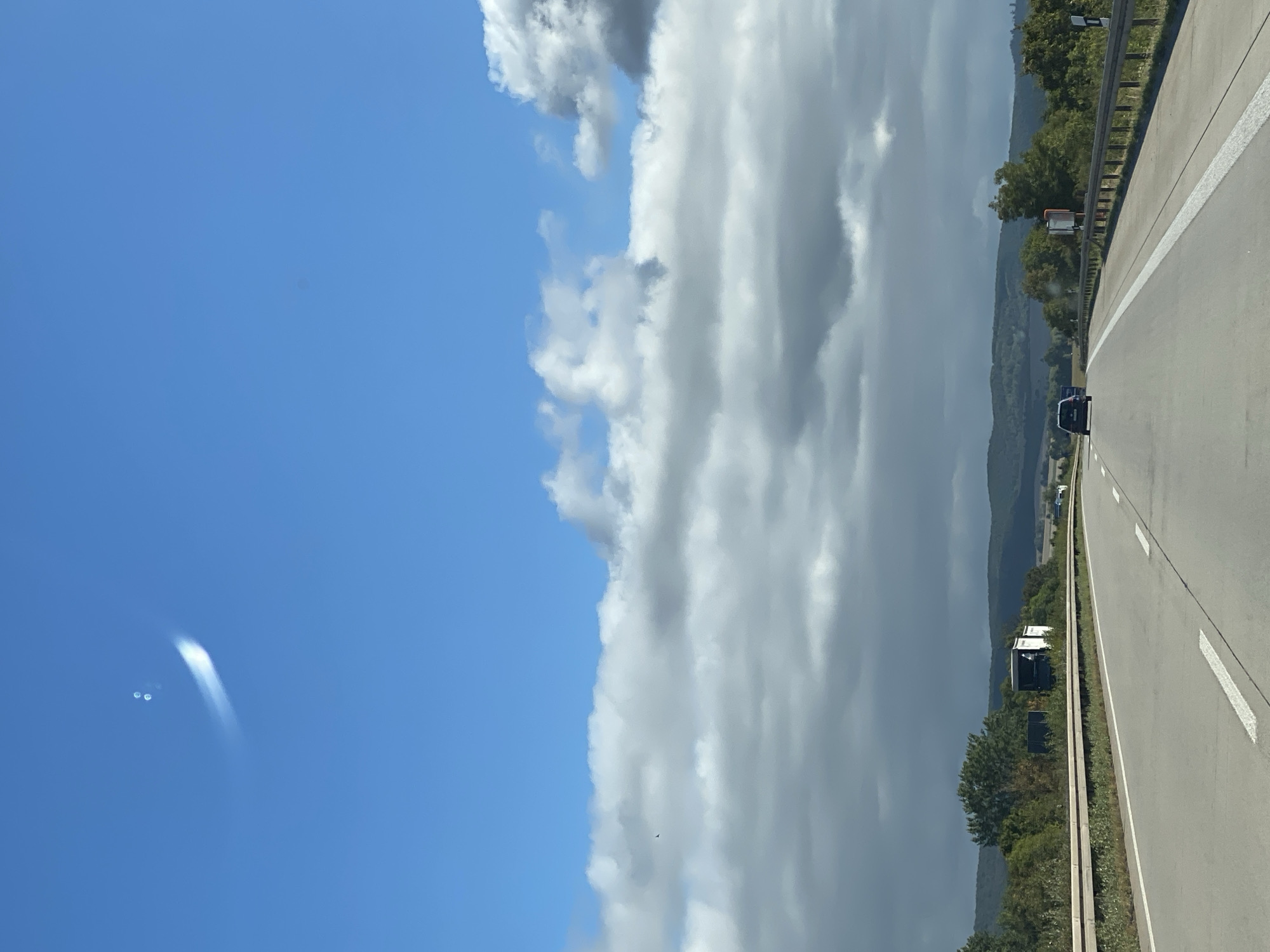 A dark blue passenger car is traveling on a multi-lane highway. The highway appears to be in a rural or semi-rural area, with trees and rolling hills visible in the distance. The sky is partly cloudy, with some cumulus clouds present. There is a white truck on the side of the road in the distance. The image is taken from the perspective of someone in a vehicle behind the blue car. The weather appears to be fair.