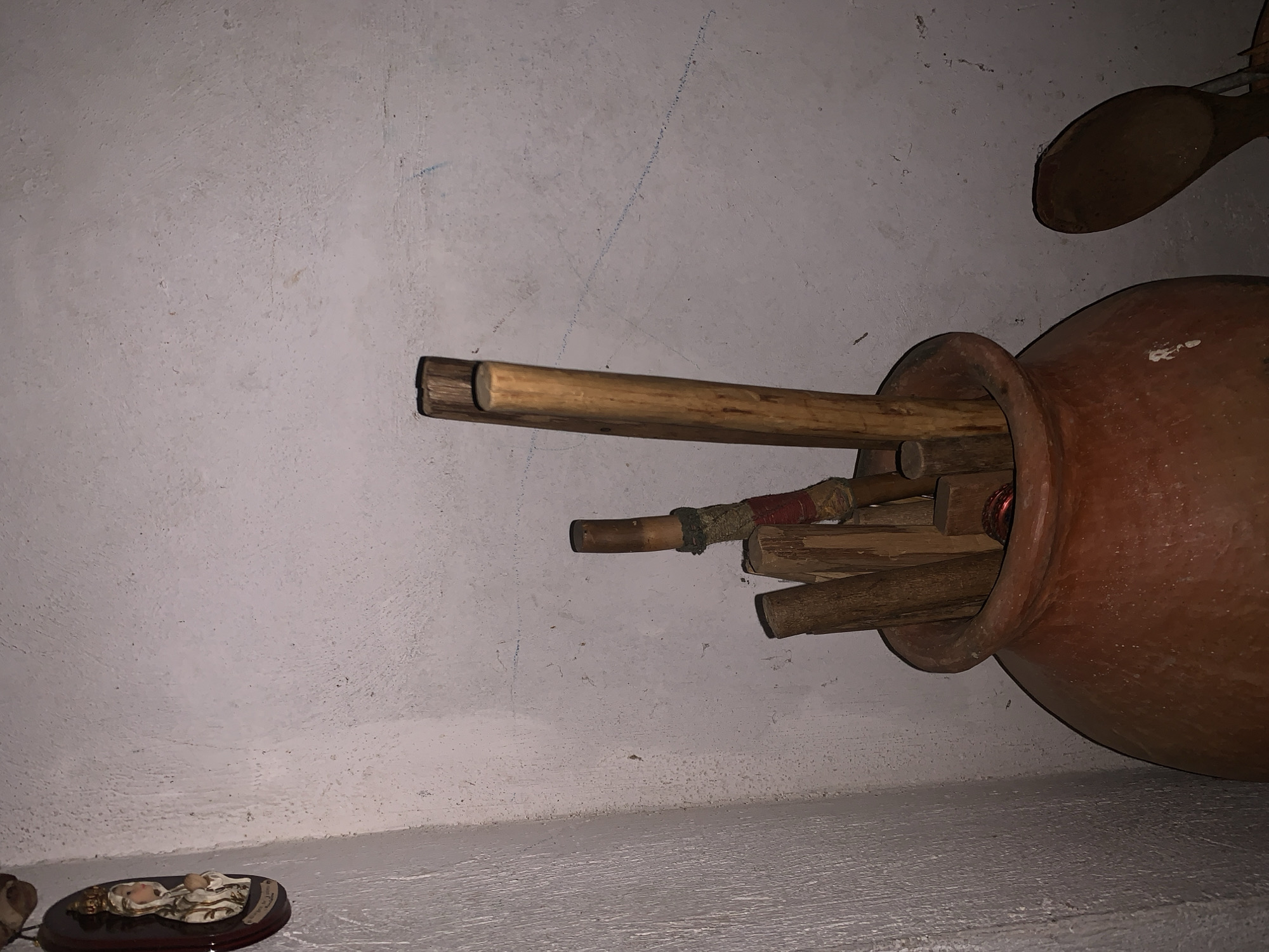 The image depicts a rustic indoor scene, possibly in Santa Ana, Venezuela, characterized by a large, reddish-brown earthenware pot lying horizontally on a light-colored shelf or surface. The pot contains an assortment of wooden sticks and dowels of varying lengths and thicknesses, some with reddish and dark cords or fabric wrapped around them. One long, light-colored wooden stick protrudes prominently from the pot.
In the upper right corner, a dark wooden spoon is partially visible, seemingly hanging or resting above the pot. On a lower surface in the bottom left of the frame, there is a small decorative plaque with a reclining figure, possibly a baby or child, resting on a dark wooden base.
The background wall is textured, appearing to be off-white or light grey stucco or concrete, showing signs of age with some faint blue markings and subtle discoloration. The lighting is dim, suggesting an indoor environment with low natural light or reliance on artificial sources. The scene is static, with no human figures or ongoing activity. There is no clearly legible text visible on any of the objects or surfaces.