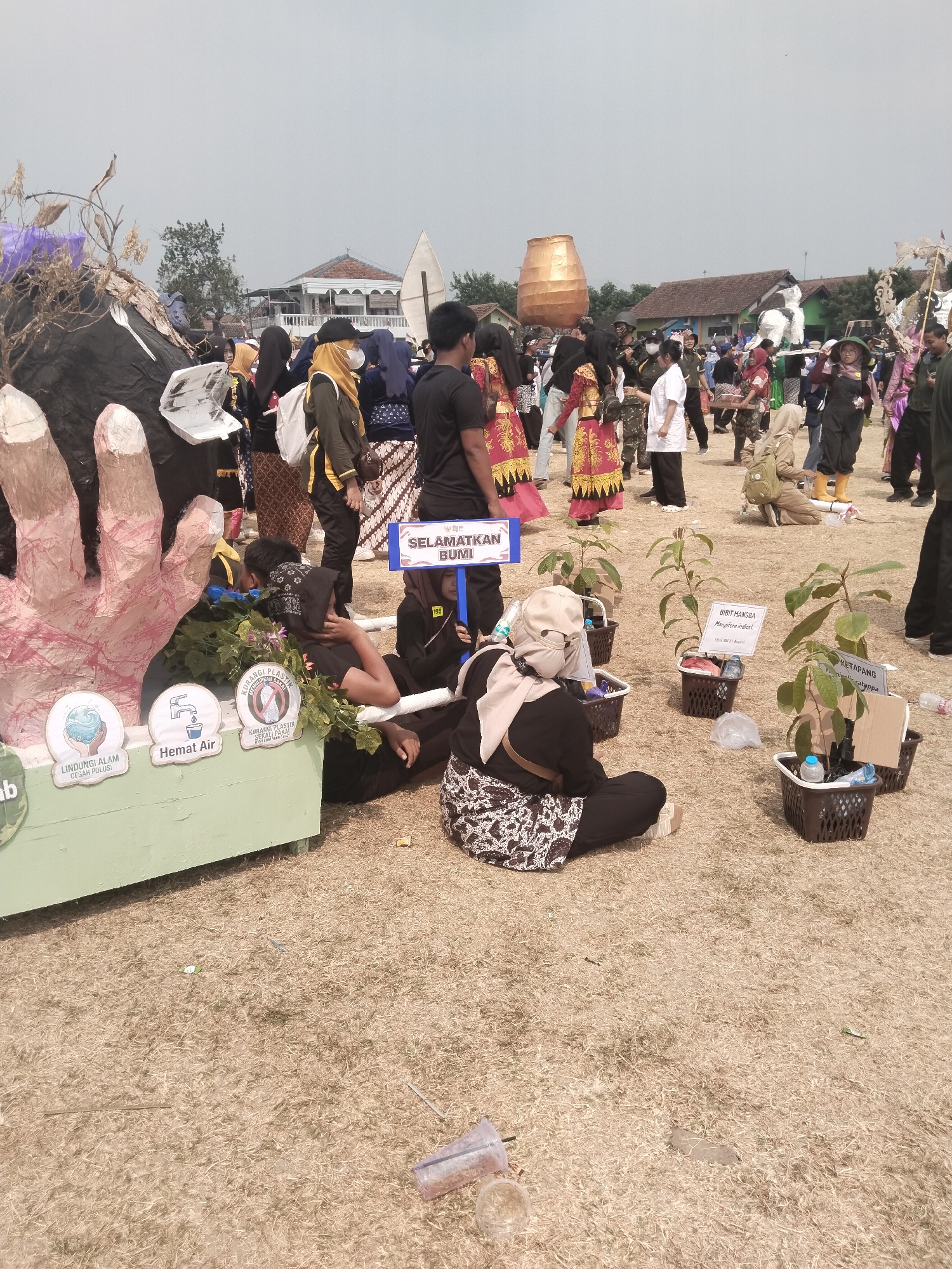 In Pakulaut, Indonesia, a public event is underway on a dry, grassy field under an overcast sky. In the foreground, a large, pastel pink and beige sculpted hand is prominently displayed, appearing to emerge from a black, amorphous structure adorned with dry branches and purple plastic. To the left of the hand, a green, wooden display board features three circular icons with text in Indonesian. The first icon depicts a swirling globe of water with the text "LINDUNGI ALAM CEMARI POLUSI" (Protect Nature from Pollution). The second icon shows a faucet with dripping water and the text "HEMAT AIR" (Save Water). The third icon displays a crossed-out plastic bottle with the text "KURANGI PLASTIK SEDINI SAATNYA" (Reduce Plastic Now is the Time).
Several individuals are situated around these displays. A person in a black headscarf and black clothing with a patterned skirt is sitting on the ground, leaning against the wooden board, partially obscured by greenery. Another person in a light-colored headscarf is also seated nearby, wearing dark clothing. Further to the right, three potted saplings are arranged in brown baskets. Each sapling has a white sign with text in Indonesian, identifying them as "BIDIK MANGGA" (Mango Seedling) and "KETAPANG" (Ketapang).
In the middle ground and background, a crowd of people, many wearing traditional Indonesian attire like batik skirts and headscarves, are gathered. Some are standing, while others appear to be walking or interacting. One individual in the center wears a yellow headscarf and dark clothing, while another in the foreground right wears a black headscarf and a patterned skirt. A blue sign with the text "SELAMATKAN BUMI" (Save the Earth) is visible amidst the crowd. The background reveals scattered buildings with pitched roofs, suggesting a semi-rural or suburban setting. The overall atmosphere suggests an environmental awareness event or festival.
