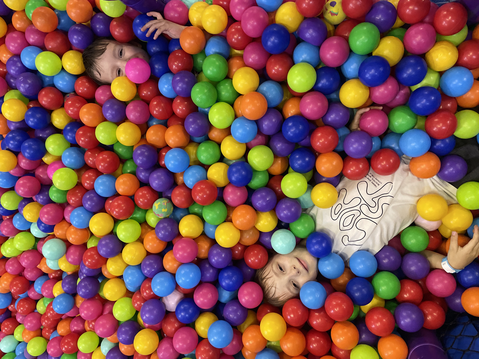 Two children are partially submerged in a ball pit containing balls of various colors, including red, yellow, green, orange, pink, purple, and blue. The first child is in the upper left, with a pink ball near their face, and a raised arm. The second child is in the lower right, wearing a white t-shirt with a black abstract design. The photograph was taken indoors. Sochi, Russia is not discernable in the image.