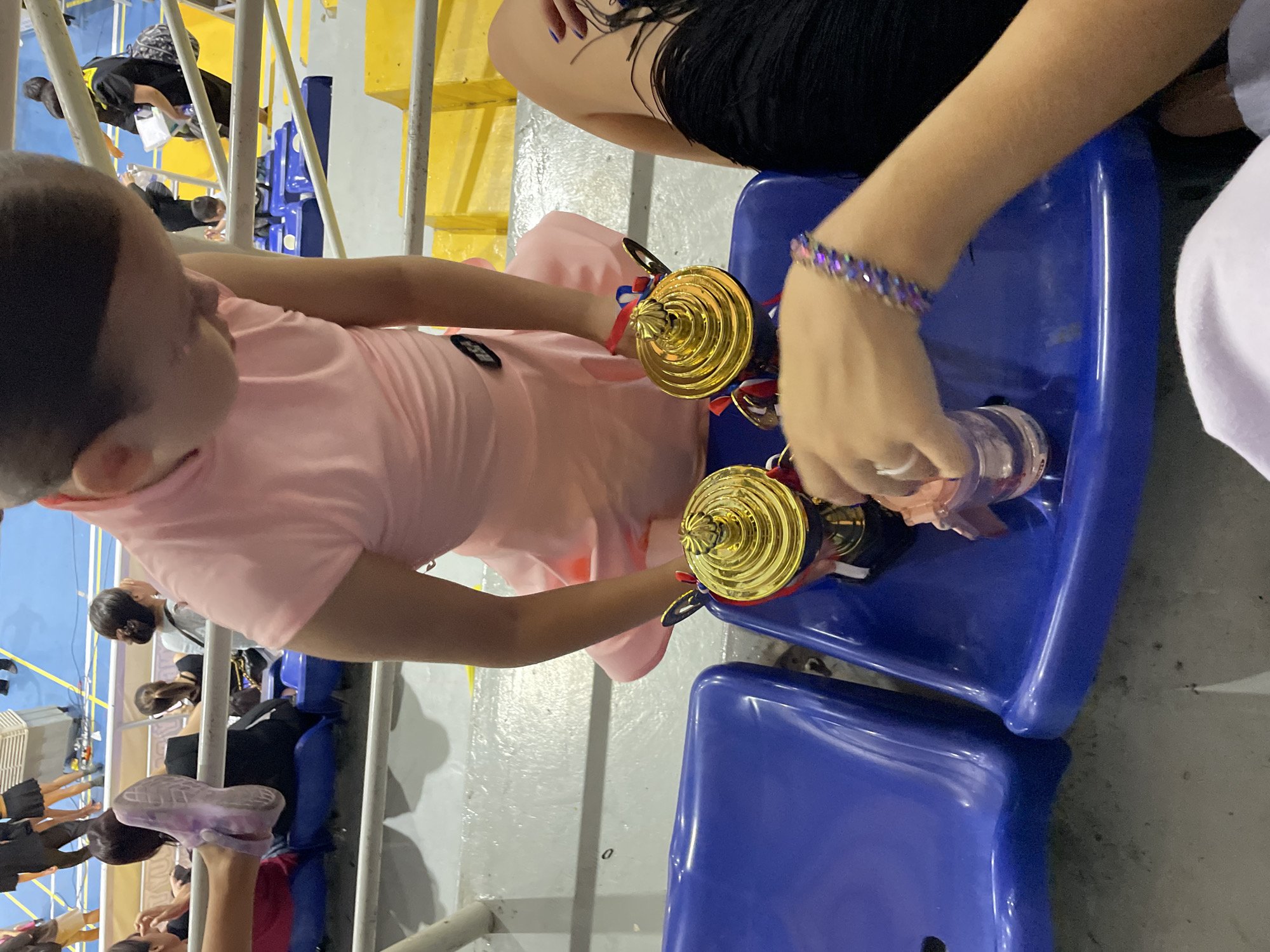 In a stadium with blue seats, two trophies are placed on a blue seat. A person in a pink dress is holding one of the trophies. Another person's arm, wearing a bracelet, is reaching for a small plastic cup next to the trophies. In the background, other people are seated, and a person in a black shirt with yellow accents is visible. The environment appears to be an indoor sports arena or event hall, possibly at a school or community center. There is no specific indication of the city of Thuận An, Vietnam within the image.