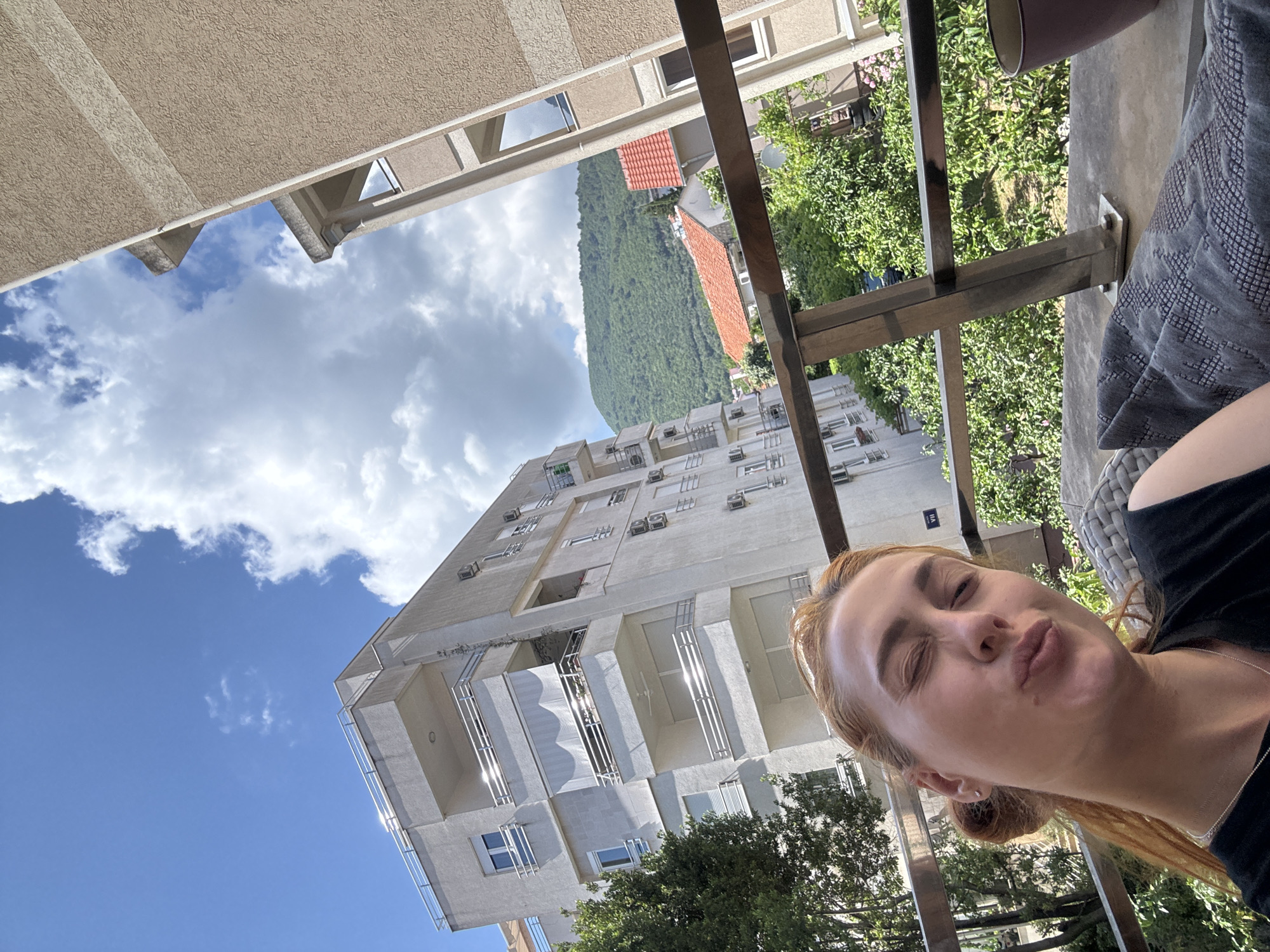 The image, originally rotated 90 degrees counter-clockwise, depicts a bright daytime scene from a balcony in Budva, Montenegro. In the foreground, on the right side of the frame (when correctly oriented), a young woman takes a selfie. She has light brown hair, pulled back in a bun, and wears a black top with a thin silver necklace and a small earring. Her eyes are slightly squinted, and her lips are pursed in a playful, "pouty" expression. Part of a grey, patterned cushion or fabric is visible next to her, along with a metal balcony railing.
Beyond the balcony, a modern, multi-story apartment building with numerous balconies and external air conditioning units stands prominently. The building is light-colored and appears to be made of concrete or stucco. Lush green trees and foliage are visible around the base of this building and in the midground. In the background, a large, verdant mountain, densely covered in trees, rises against a clear blue sky dotted with scattered white clouds. Smaller buildings with distinct red-tiled roofs are nestled at the mountain's foot. The weather is sunny and bright. No discernible text is visible within the frame.