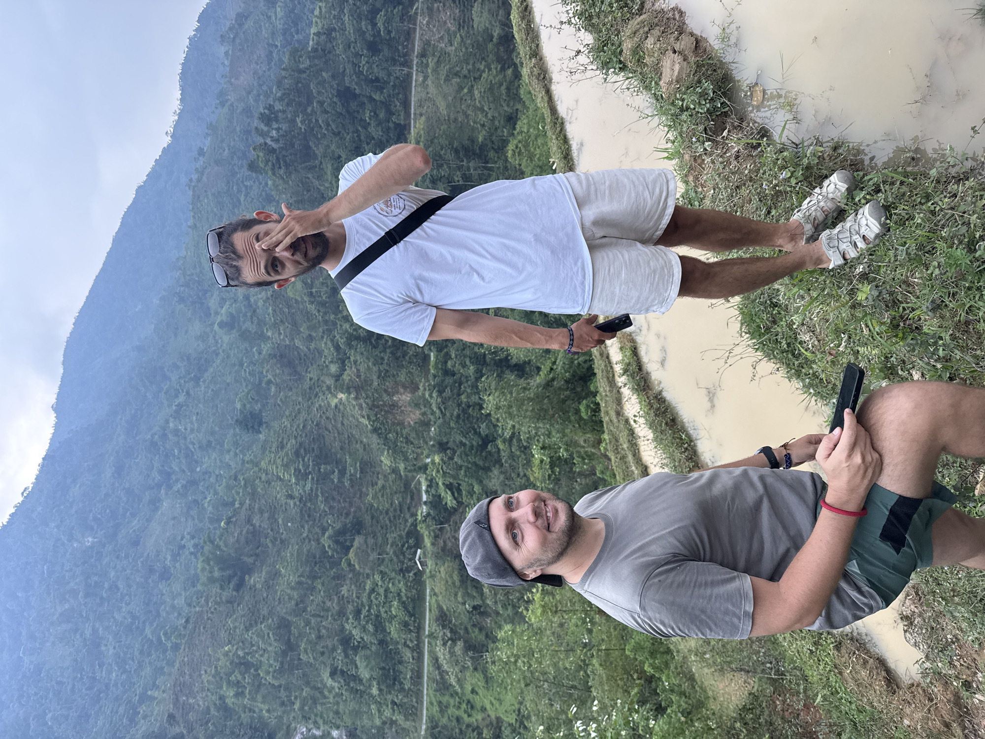 This image features two men situated in a mountainous, rural landscape within Nậm Dịch Commune, Vietnam. The scene showcases lush, forested mountains under a hazy or overcast sky, with what appear to be muddy rice paddies or irrigation channels bordering grassy banks in the foreground.
In the foreground, on the viewer's right, one man is visible. He wears a grey t-shirt, green shorts with black accents, and a grey cap. He has a beard and a subtle smile as he looks towards the left. He is holding a black smartphone, seemingly interacting with it. A red bracelet is on his right wrist, and a dark band, possibly a smartwatch, is on his left.
Further back and to the viewer's left, the second man is standing. He is dressed in a white t-shirt, light-colored shorts, and light grey sandals. Dark sunglasses rest on his head. With a beard and a slight smirk, he engages with the camera, making a playful gesture by partially covering his right eye with his hand. A black strap, likely from a bag, crosses his shoulder. He also holds a dark smartphone in his left hand.
The background consists of densely forested, steep mountains, disappearing into a soft haze. In the midground and foreground, the muddy water of the paddies or channels is visible, bordered by uneven ground with various grasses and small plants. The men's attire suggests casual outdoor activity or travel. The overall atmosphere conveys a relaxed moment during a daytime exploration of this scenic, agricultural mountain region, likely under diffused light from a cloudy sky. No discernible text is visible on any objects or clothing.