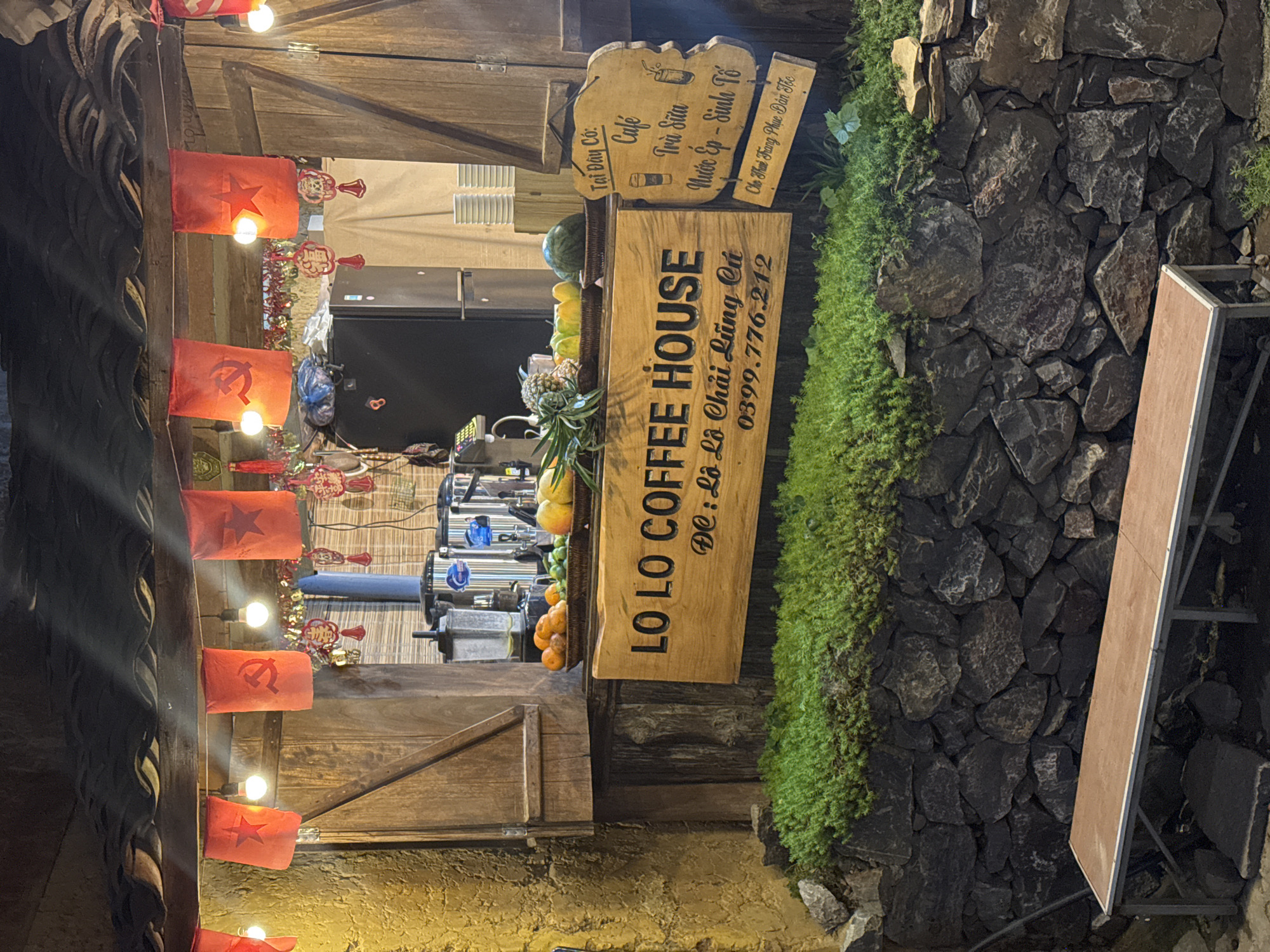 A wooden sign that reads "LO LO COFFEE HOUSE" is the most prominent object in the image. The sign also includes Vietnamese text indicating the address as Lô Lô Chải Lũng Cú, which is a commune in Vietnam, and a phone number. To the right of the sign, there's a wall made of rocks, with green moss-like vegetation growing on it. To the left of the sign, there is a display of fruits and what seems to be coffee-making equipment. Above the main sign, a smaller wooden sign lists coffee and other drinks in Vietnamese. Red flags with stars, along with lights, hang from the top of the image, likely part of a decorative feature. The flags may be indicative of Vietnamese national symbolism. The image is taken at night.