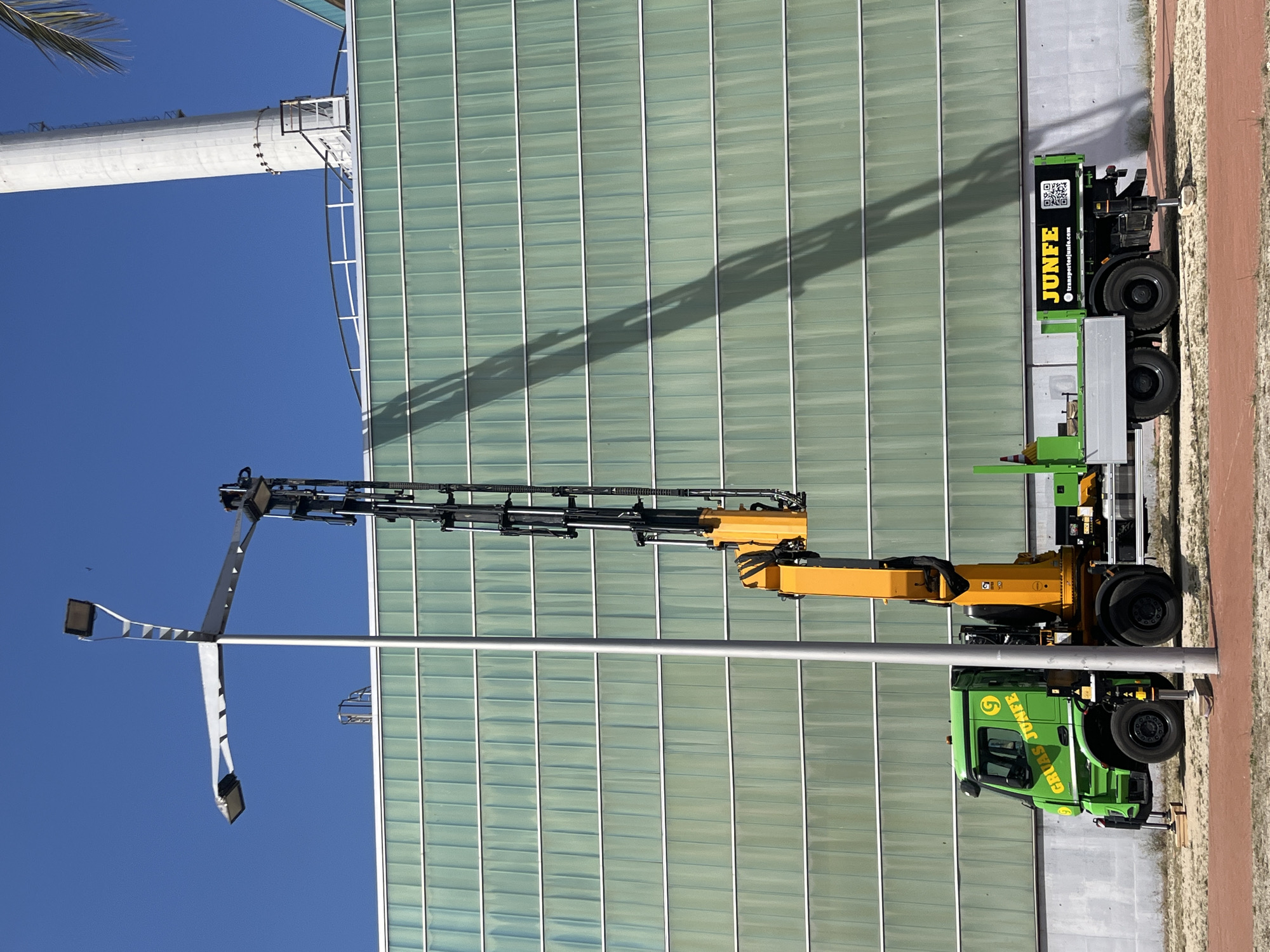 A bright green truck with a yellow crane is parked on a sandy surface with a red-colored path in the foreground. The truck is branded with "GRÚAS JUNFE" on the side of the cab and "JUNFE" with a QR code and website address on the rear. The crane is extended vertically, casting a long shadow on the glass facade of a modern building behind it. The building has horizontal green glass panels and is adjacent to a tall, white cylindrical structure, possibly a chimney or industrial tower. A street lamp with multiple lights is also visible to the left of the truck. The sky is a clear, vibrant blue. The location is identified as Sant Adrià de Besòs, Spain, likely near an industrial or commercial area. There are no people visible in the image.
