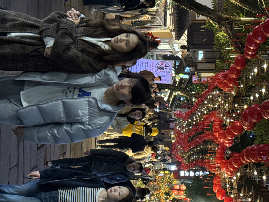 This image captures a nighttime scene on a bustling pedestrian street in Yuzhong District, China. The street is densely populated with individuals, primarily of East Asian descent, walking in various directions. Hundreds of decorative red lanterns, many with tassels, are strung in multiple rows overhead, forming a canopy across the street. These lanterns are interspersed with strings of small white electric lights, illuminating the area.
Traditional-style buildings with dark tiled roofs and illuminated storefronts line both sides of the cobblestone-paved street. Several signs in Chinese characters are visible; one prominent sign on the left features a white background with red characters reading "小屋" (Xiǎo wū), along with details that appear to be operating hours. Trees with visible branches are present along the street, integrated with the overhead lighting structures. In the mid-ground on the right, a plant bearing small, round, orange fruits is illuminated. The street slopes gently uphill towards the background.
In the immediate foreground, a woman and a man walk side-by-side towards the left side of the frame. The woman, positioned slightly ahead and to the left, has long, dark hair and is wearing a brown and beige plaid long coat over a light-colored top, with her hands clasped in front. The man, walking next to her, has short, dark hair and wears a light grey puffer jacket over a light grey t-shirt featuring a blue graphic design and a Nike swoosh logo. He wears grey pants.
Further back in the crowd, a person in a bright yellow uniform or jacket with black markings on the back is visible, walking away from the camera. Other individuals in the mid-ground and background are dressed in casual winter attire, navigating the crowded thoroughfare. The overall scene depicts a vibrant, festive atmosphere.