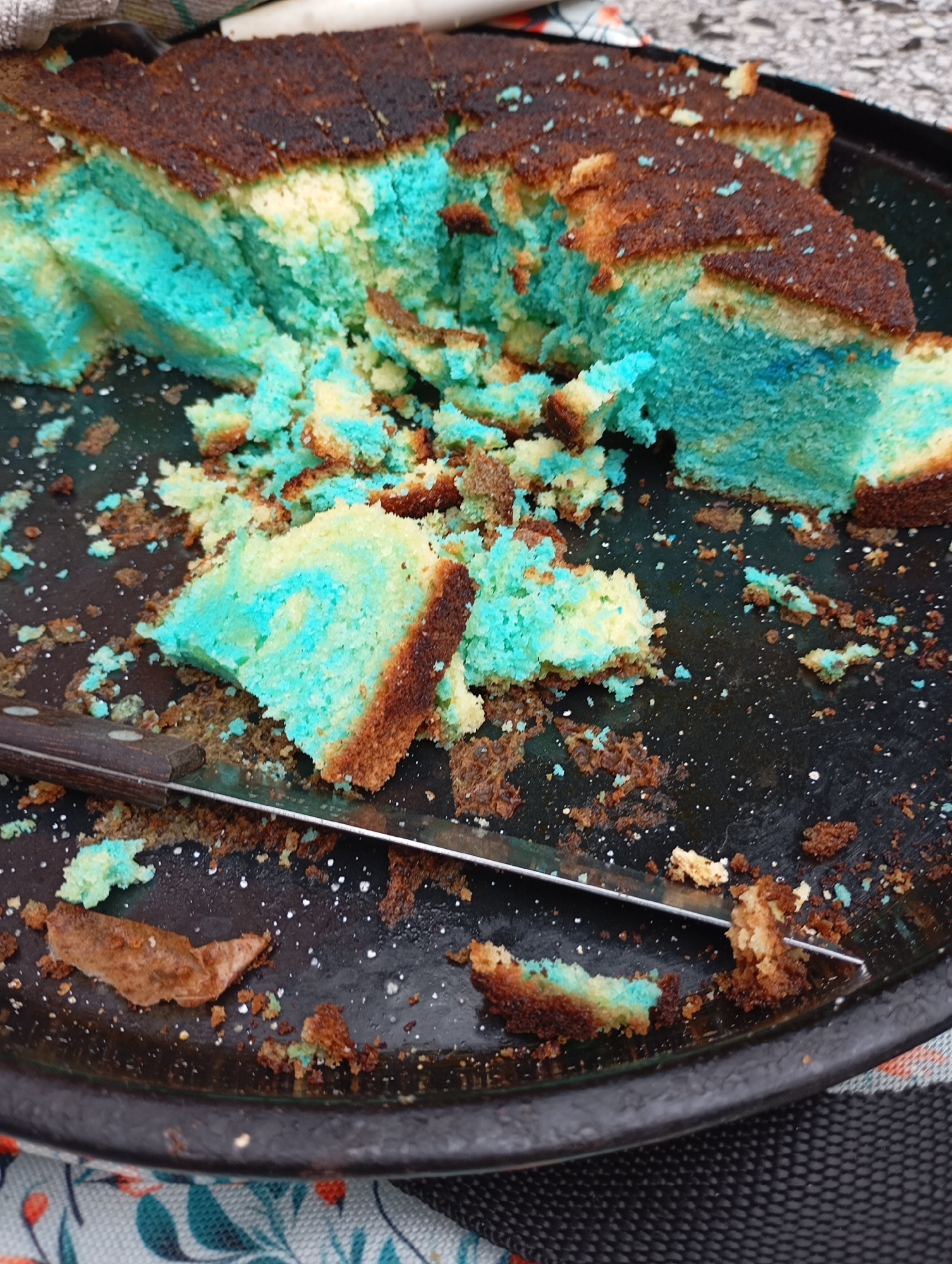 A close-up, top-down view captures a partially eaten cake with a marbled pattern of blue and yellow cake layers on a dark, circular platter. The cake is cut into several pieces, with many crumbs scattered around the platter and on the cake itself. A silver knife with a dark brown handle rests across the cake and platter. The platter is placed on a surface covered with a patterned cloth and a textured black mat. The image does not provide enough information to determine its location in the city of La Plata, Argentina.