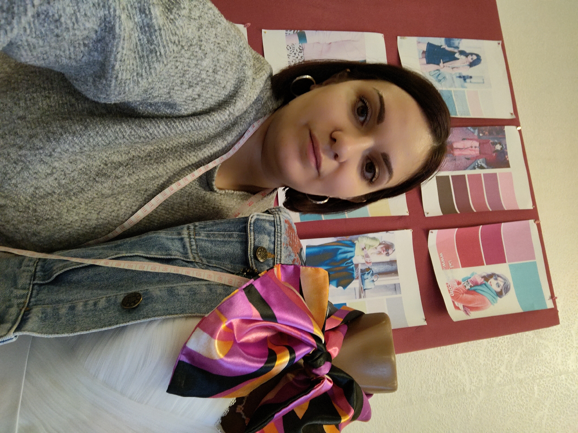 A young woman with dark hair and a neutral expression looks directly at the camera in a close-up, selfie-style shot. She is wearing a light grey, textured sweater and large hoop earrings. A pink and white measuring tape is draped around her neck and shoulder, with numbers like "108" through "115" and "25" visible on it.
To her left, a denim jacket is partially visible, and below it, a mannequin bust displays a vibrant, multi-colored satin scarf tied in a bow. The scarf features bold patterns in black, pink, orange, yellow, and purple.
The background consists of a reddish-brown board pinned with several sheets of paper. These appear to be fashion design mood boards or inspiration sheets. One sheet shows an illustration of a woman in a black dress with an accompanying blue, white, and grey color palette. Another displays an image of a person in red attire with a pink, red, and brown color scheme. A third sheet features an illustration of a woman in an orange jacket and a turquoise scarf, paired with a similar color palette, and partially visible text that reads "www.a..." (likely part of a website address).
The setting suggests a creative space, possibly a design studio or workshop, emphasizing fashion or textile work, given the presence of the measuring tape, mannequin, and design boards. The lighting indicates it is daytime.