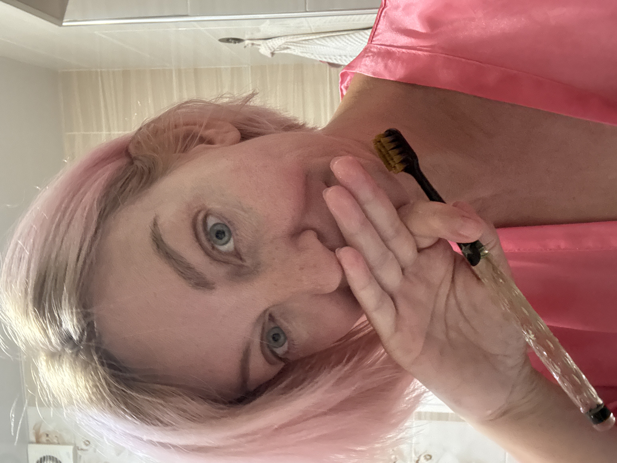 A woman with short, pink-dyed hair is taking a selfie in a bathroom. She is wearing a pink satin robe and is holding a toothbrush with a clear, textured handle and a black toothbrush head with yellow bristles. Her right hand is covering her mouth, and she is looking at the camera with a slight smile. The background shows a tiled wall, a light fixture, and a towel hanging on a hook. The scene appears to be in a domestic bathroom, not specifically identifiable as being in Sochi, Russia.