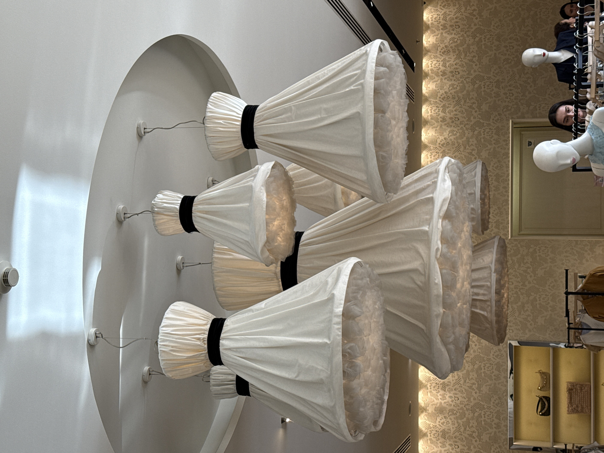The image shows an interior space with a decorative lighting fixture dominating the left side. This fixture features a cluster of white, pleated lampshades, each cinched with a black band, mounted on a rounded white surface.
On the right side of the frame, there are racks of clothing and mannequins visible in the background. A patterned wallpaper can be seen behind the mannequins.
The location is Moscow, Russia.