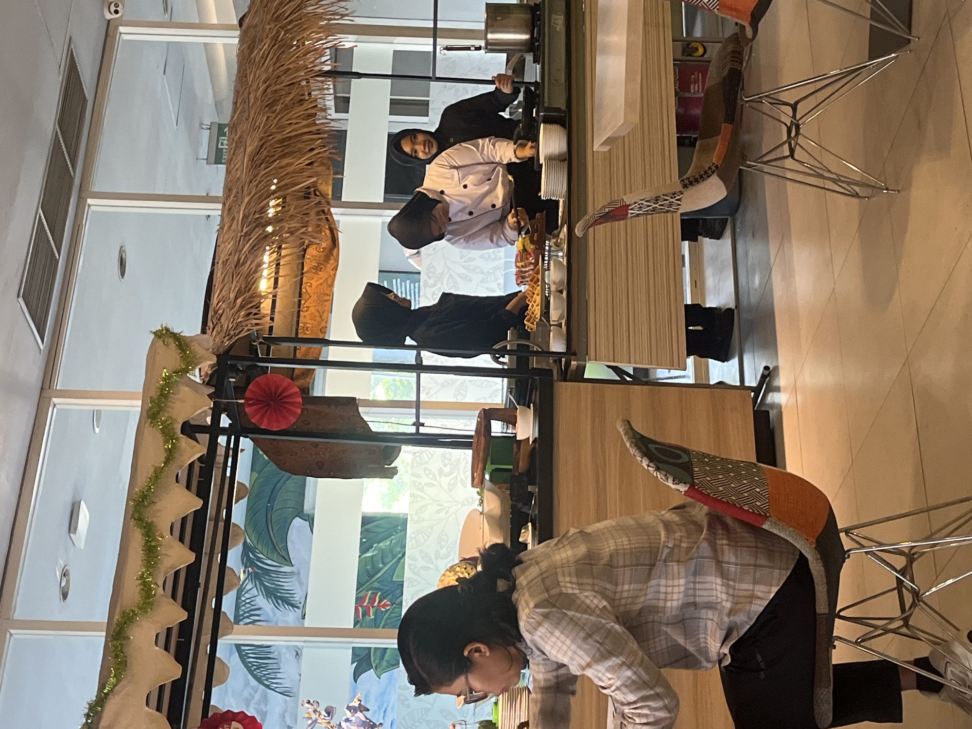 The image depicts a buffet-style food service area with a thatched roof, resembling a tropical-themed setup, likely in Gapurosukolilo, Indonesia. In the foreground, a person wearing glasses and a plaid shirt is seated at a table, facing away from the camera. Behind them, three individuals in black headscarves are interacting with food. One person, dressed in a chef's uniform, is actively cooking. To the left, a display features waffles and other food items. In the background, large windows offer a view of lush green foliage, possibly a mural, with palm trees and vibrant flowers. The overall environment suggests a restaurant or catering facility with an emphasis on a relaxed, perhaps beach-resort-like atmosphere.