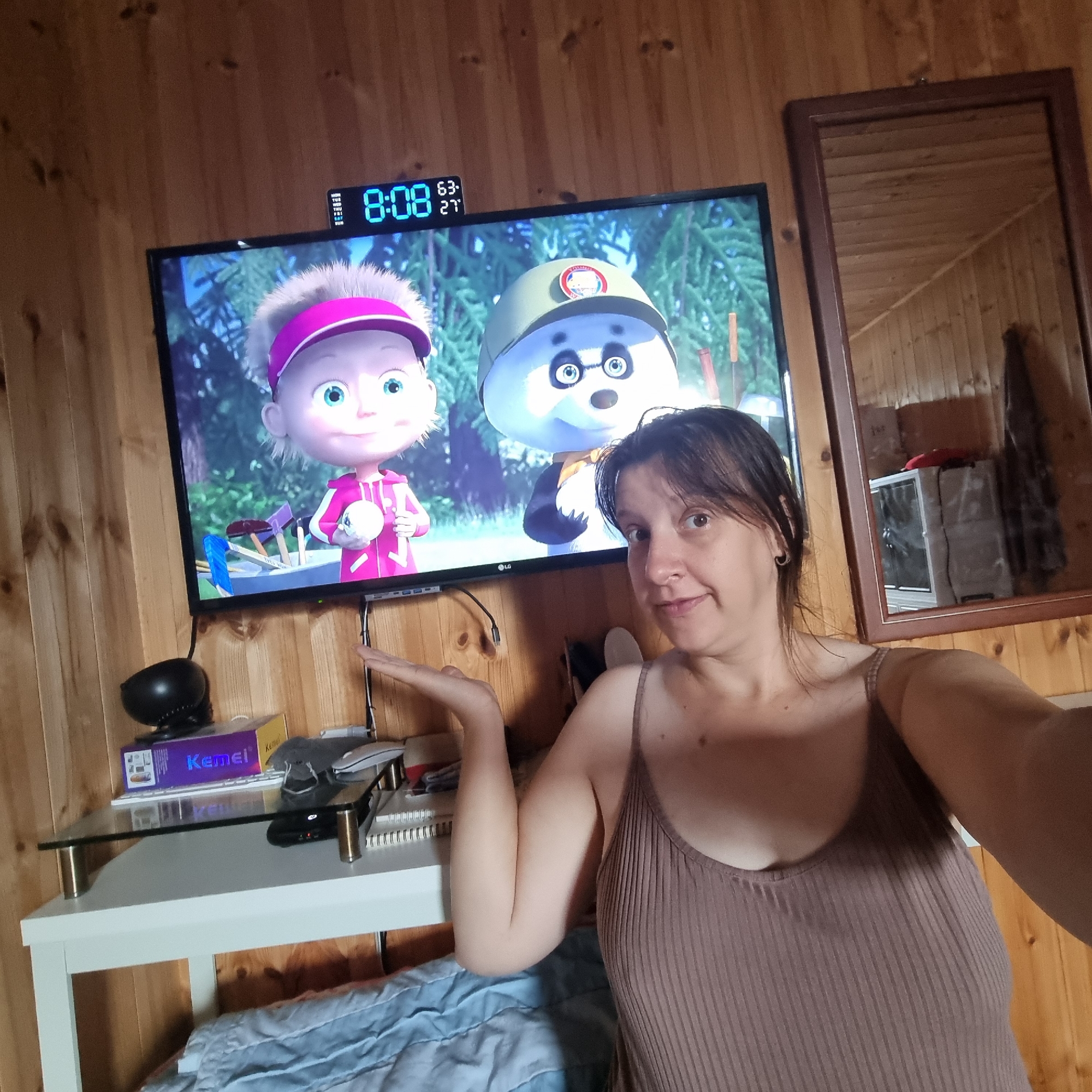 A medium shot captures a woman in a tan ribbed tank top sitting in front of a television playing an animated show featuring two characters. The woman's left hand is raised, palm facing outwards, in a gesture that could be interpreted as presenting or questioning. She is looking towards the camera with a slight smile. The television screen displays the characters Masha and the Bear, specifically Masha with a pink visor and a panda wearing a helmet. Above the TV, a digital clock displays "8:08" along with temperature readings of "63" and "27". The environment appears to be a room with wood-paneled walls. To the right of the television, a wooden-framed mirror reflects a portion of the room, including what appears to be storage units. In the foreground, on a white table with a glass shelf, are various electronic devices, including a black speaker, a box labeled "Kemei", a white keyboard, and a computer mouse. A blue blanket is visible on a surface in the lower part of the image. The location is not specified as Dunnae-myeon, South Korea.
