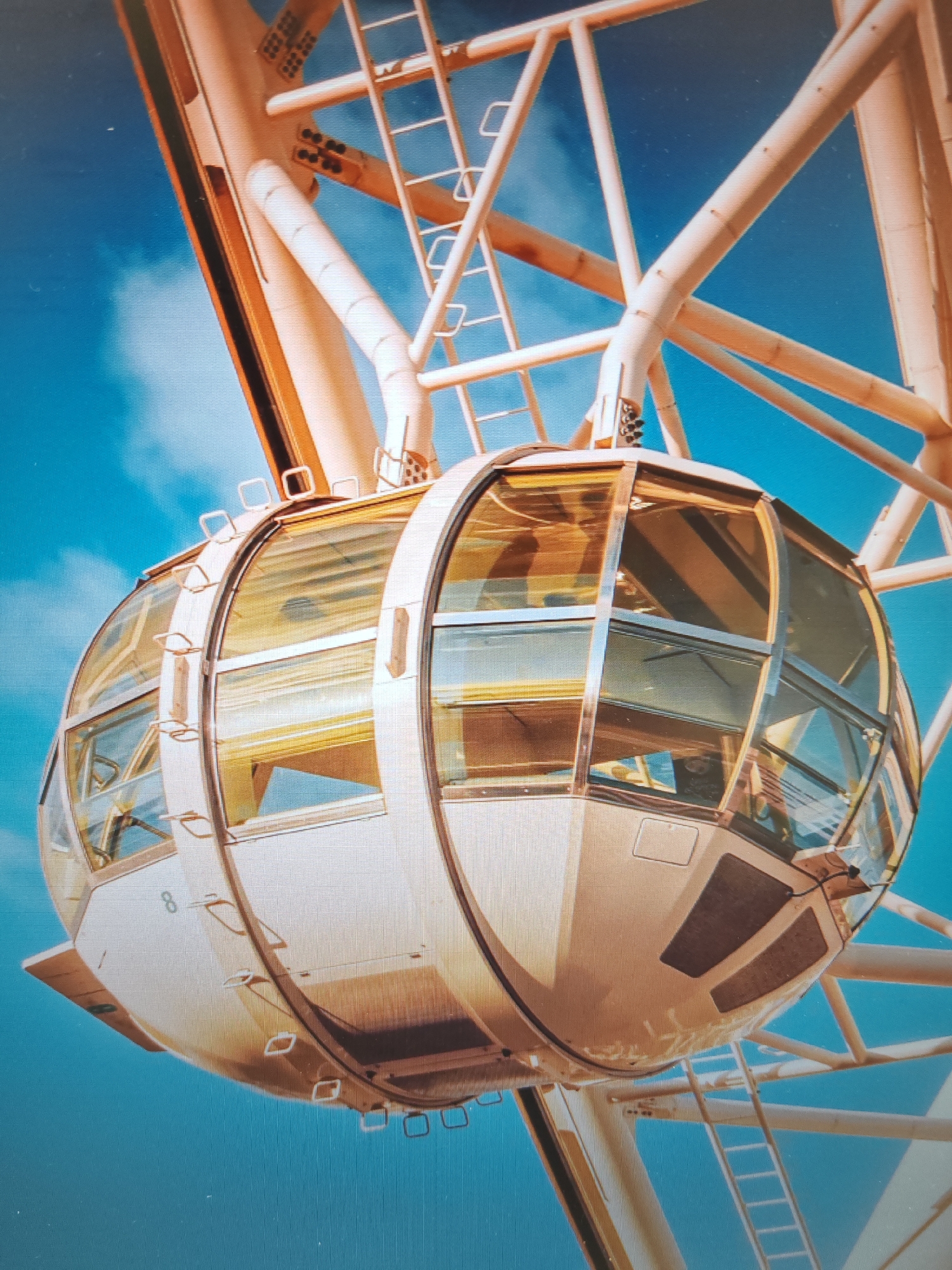 The image shows a close-up of a gondola on a Ferris wheel against a bright blue sky with some clouds. The gondola is oval-shaped with large glass windows framed by a cream-colored structure. Inside the gondola, yellow lighting is visible, reflecting off the glass. The Ferris wheel's cream-colored metal framework, including beams and a ladder, is also visible, extending upwards and to the sides. The gondola has the number "8" subtly visible on its exterior. This Ferris wheel is located in the city of Plovdiv, Bulgaria, and is a prominent feature of the city's skyline.