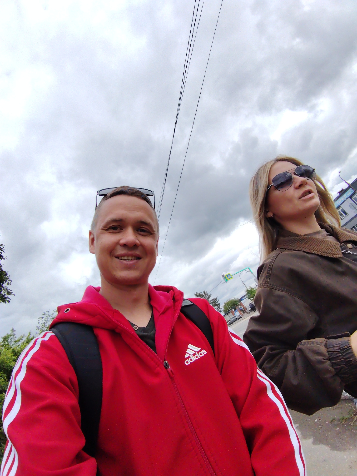 A selfie-style image shows two people, a man and a woman, outdoors on a cloudy day. The man is closer to the camera, wearing a red Adidas jacket with white stripes and a black backpack. Sunglasses are perched on top of his head, and he is smiling. The woman is slightly behind and to the right of the man, wearing sunglasses and a brown jacket.
In the background, there is a street scene with trees, a traffic light, and a building. Based on these details, it is likely that the location is the city of Chaikovsky, Russia. There are overhead power lines in the upper part of the frame.