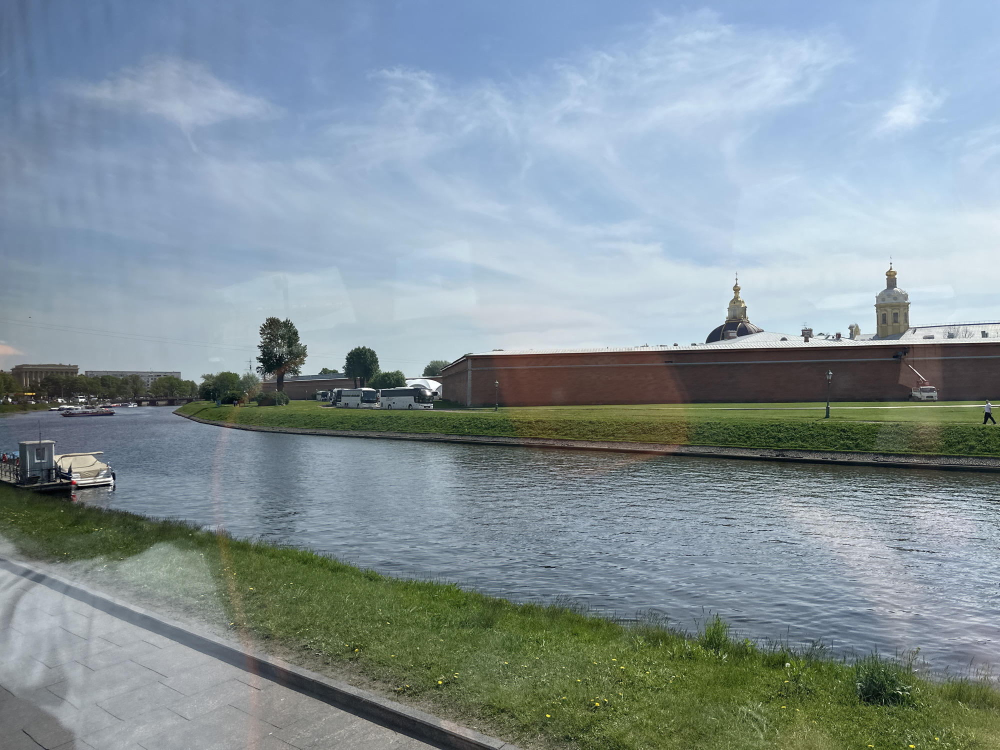 The image depicts a view of the Neva River canal in Saint Petersburg, Russia, likely taken from a moving vehicle. The foreground features a grassy riverbank with a concrete walkway visible at the bottom. A boat dock with a small white boat is visible in the water. Across the canal, a red brick wall forms the perimeter of the Peter and Paul Fortress, with a grassy area adjacent to the wall where several tour buses are parked. The spires of the Peter and Paul Cathedral rise above the wall. In the background, buildings are visible along the canal, and the sky is partly cloudy. A person is seen walking along the canal path on the far side.