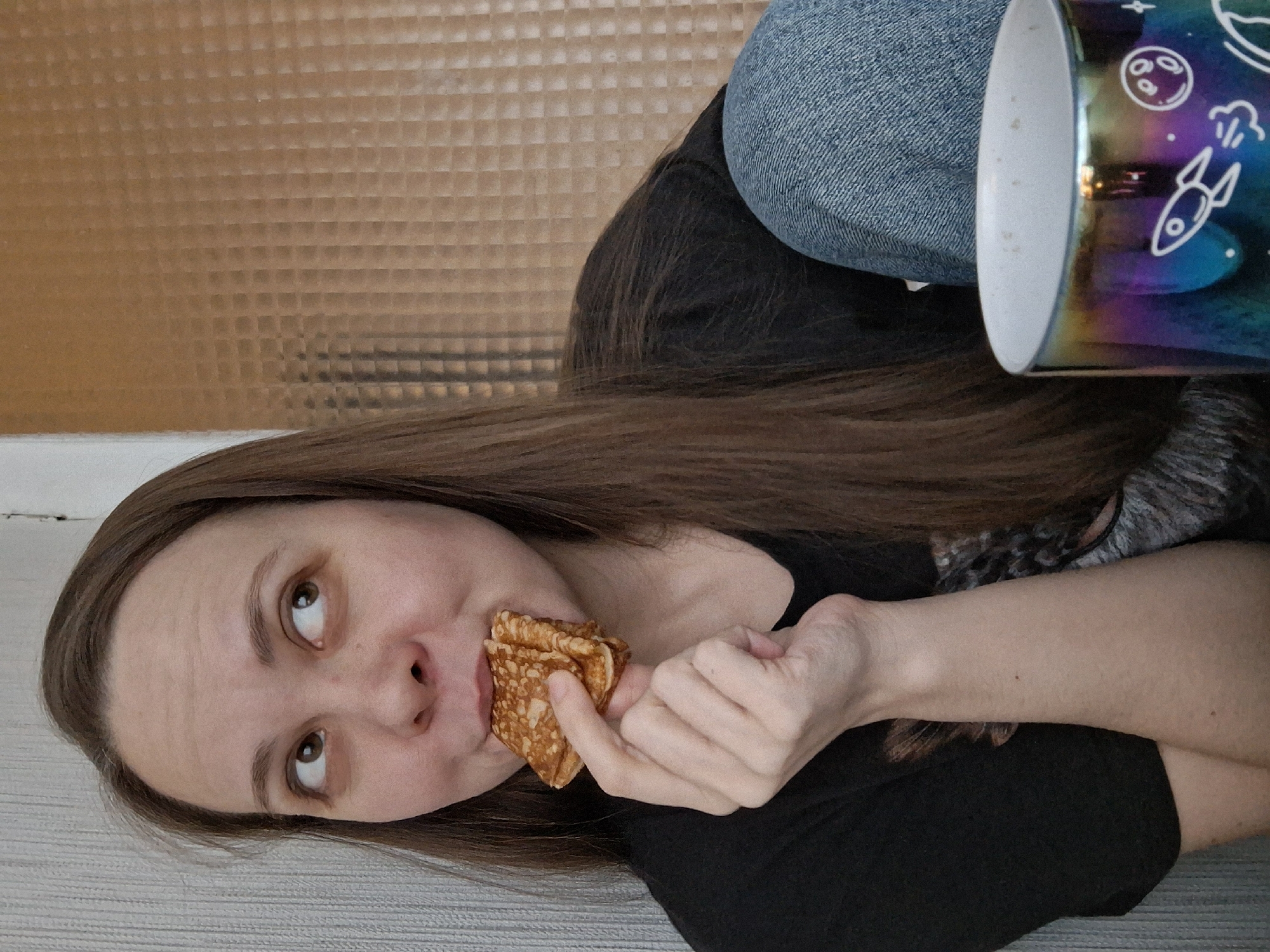 A woman is eating a crepe. She is holding the crepe in her right hand, about to take a bite. She is wearing a black t-shirt and blue jeans. There is a mug with space-themed decorations on it next to her. The location is Chaikovsky, Russia. The background appears to be a textured wall. The floor is grey.