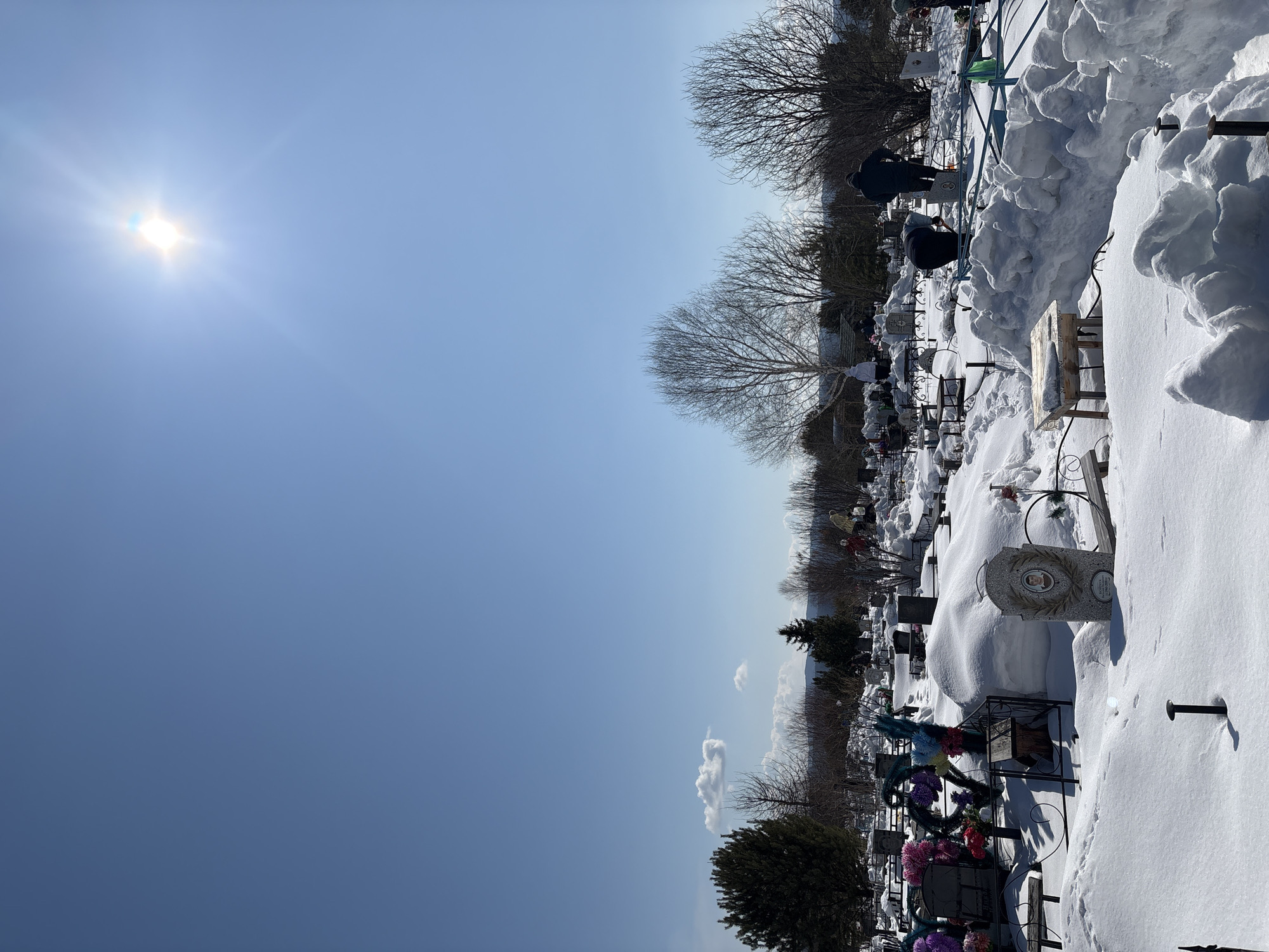 This image depicts a snow-covered cemetery in Невон, Russia, under a bright, clear blue sky. The scene is dominated by deep, untouched white snow blanketing the ground and piling up around numerous gravestones and memorials.
The sun shines intensely in the upper left, creating a prominent lens flare and illuminating the crisp winter day. The sky is a vibrant blue, with a few small, scattered clouds visible towards the horizon. In the mid-ground and background, bare deciduous trees stand stark against the sky, their branches devoid of leaves, further emphasizing the winter setting. Some evergreen shrubs are also visible.
Scattered throughout the cemetery are multiple gravestones and memorial markers. One prominent headstone in the foreground features an oval portrait of a person and what appears to be Cyrillic text with dates inscribed below. Many graves are adorned with colorful artificial floral arrangements and wreaths, adding bursts of color against the monochromatic snow. Some plots are surrounded by decorative metal fences or structures, including one with a green metal frame over it in the mid-ground.
Several people, dressed in dark winter attire, are visible among the graves. They appear to be visiting or tending to the burial sites, with one individual seen bent over near a grave, suggesting an act of remembrance or care. The clear weather and bright sunshine indicate it is daytime, likely mid-morning or afternoon. The scene conveys a sense of peaceful remembrance despite the cold, snowy conditions.