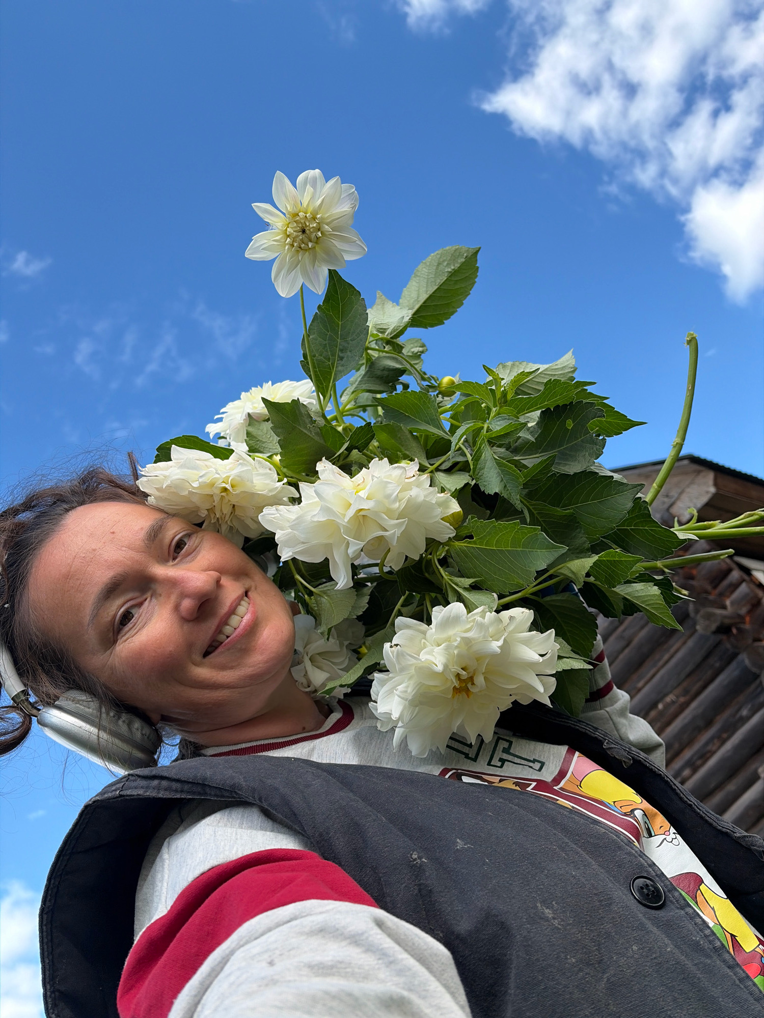 A woman with dark hair, wearing headphones, a grey sweatshirt with red trim, and a dark vest, smiles while holding a bouquet of white dahlias in front of a blue sky with scattered clouds. A wooden structure is visible in the background. The image appears to be taken outdoors, possibly in a garden or rural area, which aligns with the general location of Olkhovka, Russia.