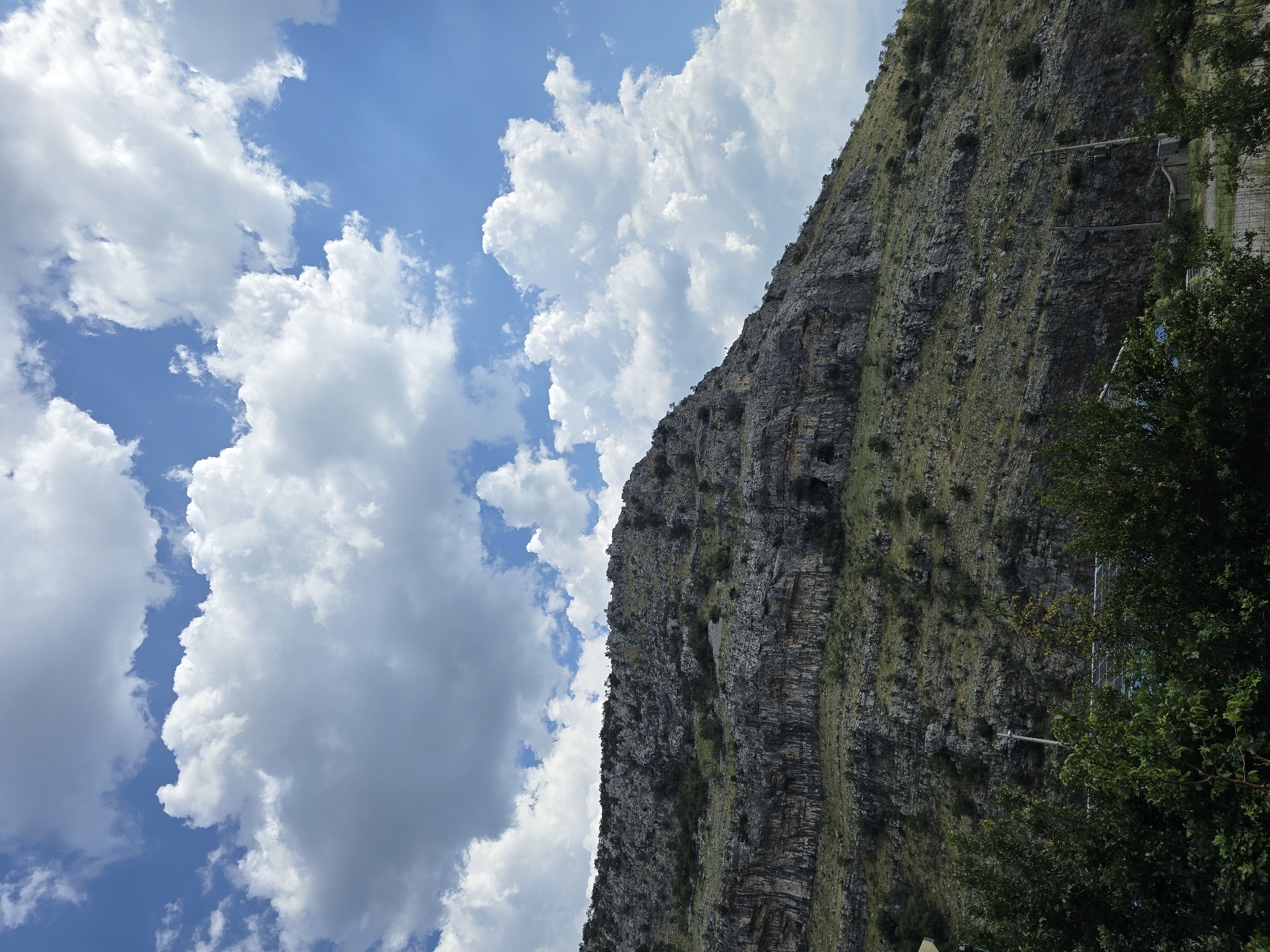 The image shows a large, rocky mountain rising from the landscape under a partly cloudy blue sky. The mountain's face is rough and textured, with visible layers of rock and sparse vegetation clinging to its slopes. Several small dark openings, possibly caves, are visible in the rock face. The foreground features dense green foliage, with some trees and bushes. Power lines and a utility pole are present along the base of the mountain, suggesting proximity to infrastructure. The scene appears to be a natural landscape with some human elements. This landscape is characteristic of the area around Podgorica, Montenegro, which is known for its mountainous terrain and proximity to natural attractions.