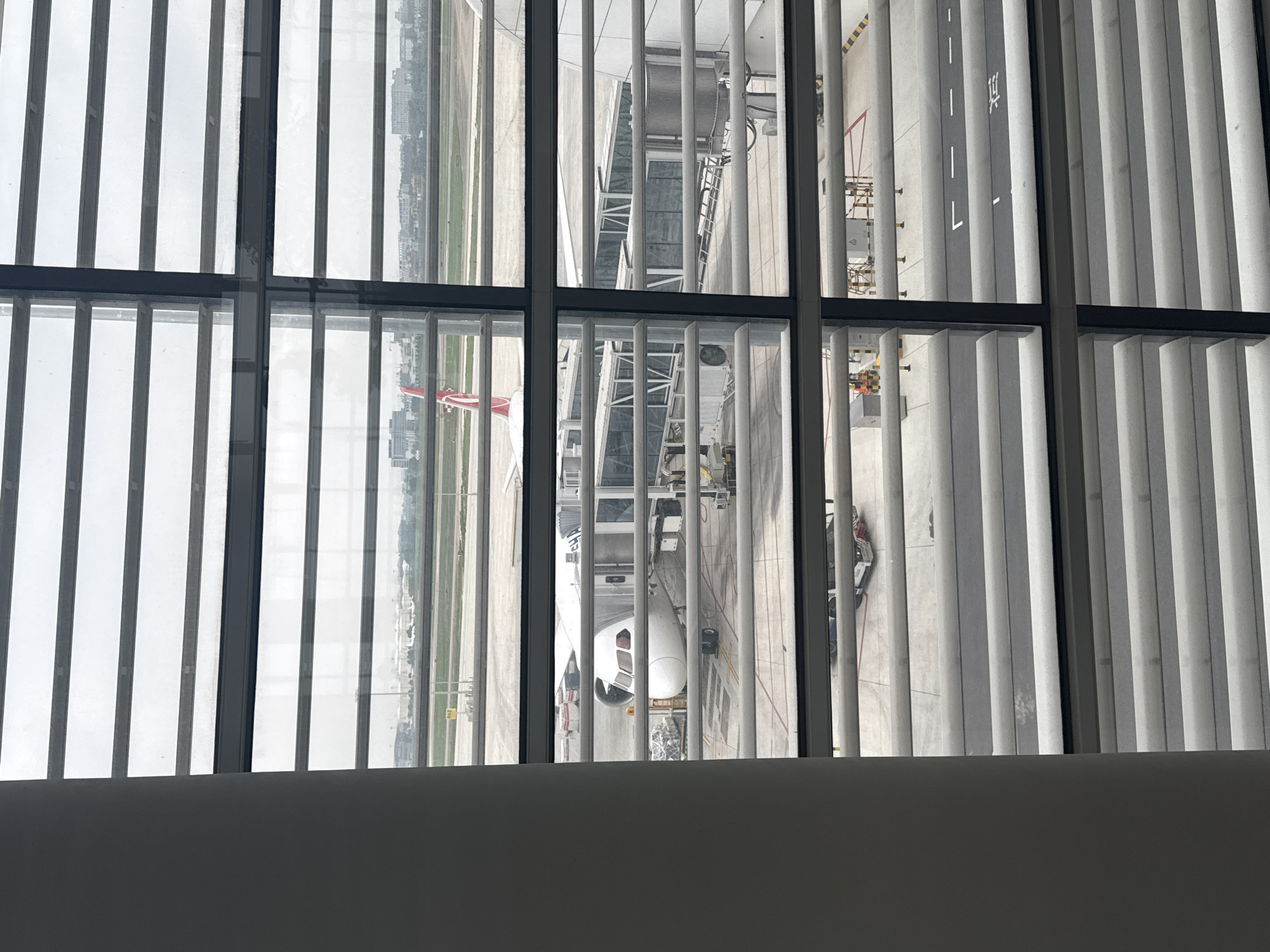 An airplane is viewed from inside a building, possibly an airport terminal, through a window with vertical blinds. The airplane is connected to a boarding bridge. Outside the window is an airport tarmac with painted markings and service vehicles. A portion of a city can be seen in the distance, and given the query parameters, the location is likely in Baiyun District, China.