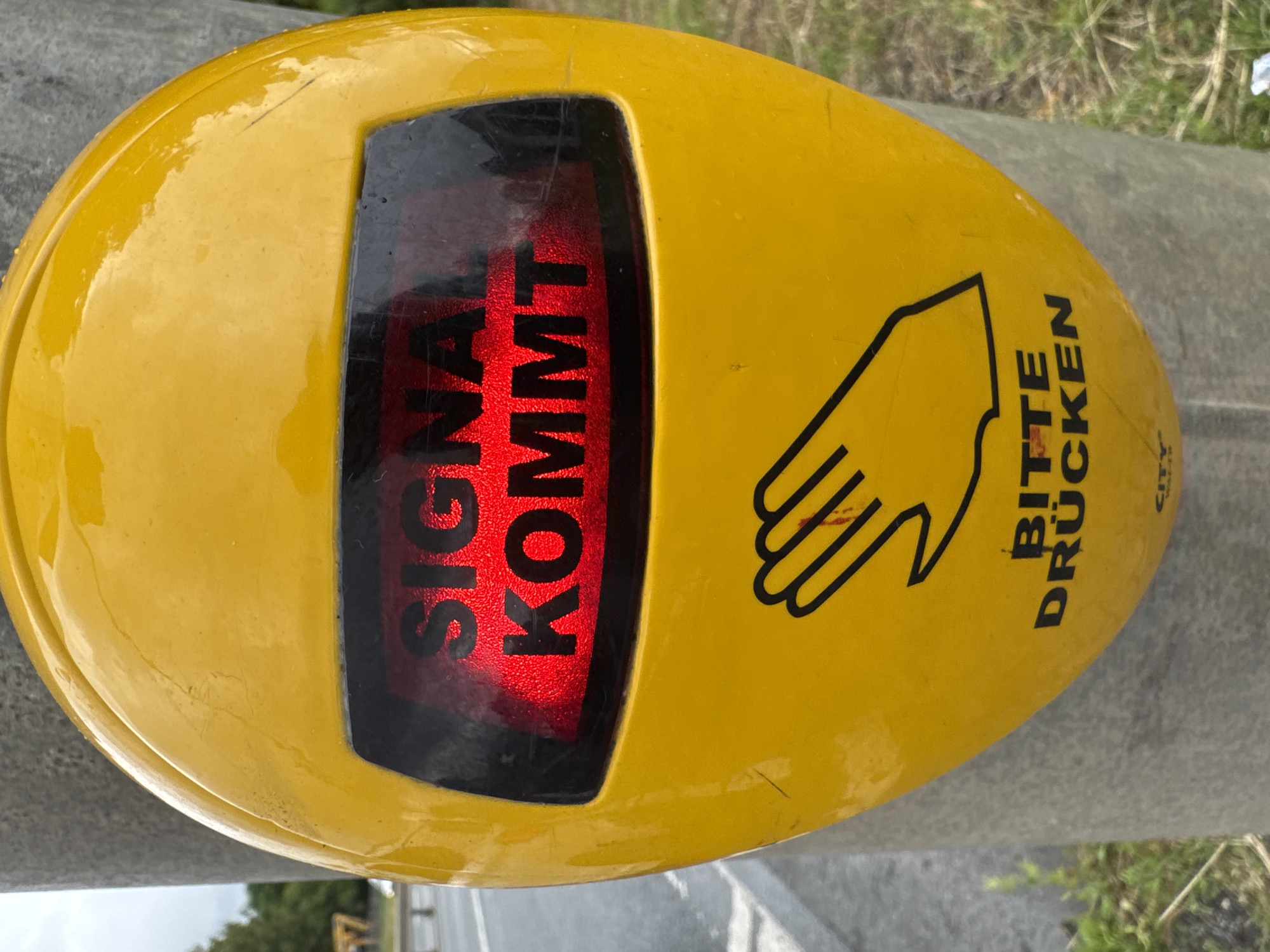 A close-up, eye-level shot features a bright yellow pedestrian crossing signal box mounted on a concrete pole. The signal box has a red illuminated display showing the German words "SIGNAL KOMMT" in bold black lettering. Below the display, a black outline of an open hand is visible, with the German words "BITTE DRÜCKEN" (Please Press) printed beneath it. The background is blurred, showing a road and some greenery, suggesting an outdoor urban environment. The image is sharp and clear, with good lighting.