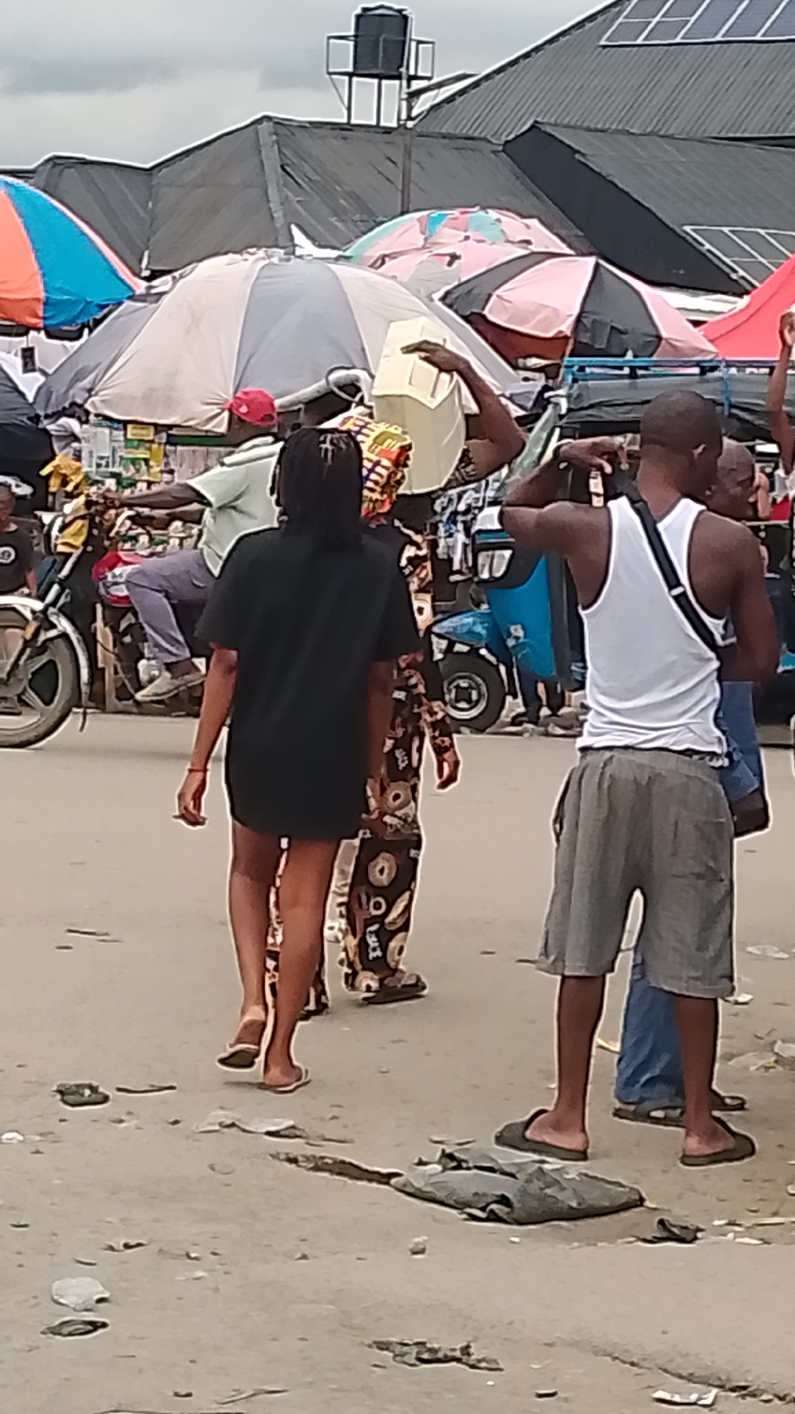 The image shows a street scene in Ukan, Nigeria. In the foreground, a woman wearing a short black dress and flip-flops walks away from the camera. Beside her is a woman in a patterned dress. To the right, a man in a white tank top and grey shorts stands facing away from the camera. He is wearing sandals and has a bag slung across his chest. In the background, there are several people, motorcycles, and a tricycle taxi. Colorful market umbrellas dot the scene. A water tower is visible above the rooftops. Trash litters the street. Solar panels are visible on the rooftops in the background.