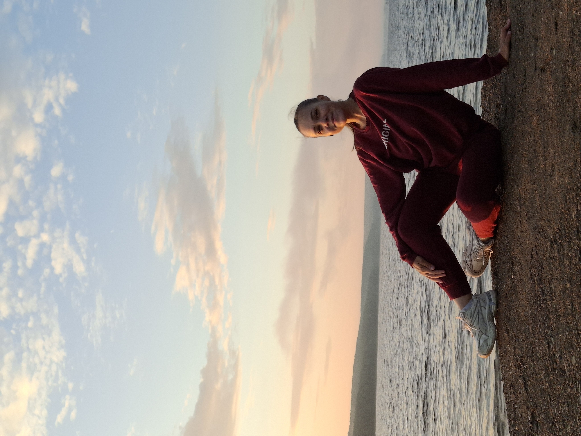 A young woman with her hair pulled back is sitting on a rocky shore, facing the camera and smiling. She is wearing a maroon sweatshirt with white lettering and matching sweatpants, and white sneakers. Her legs are crossed, with her left leg bent at the knee and her right leg extended. The sun is setting, casting a warm glow over the scene and painting the sky with shades of pink, orange, and blue. A calm body of water, likely the Kama River, stretches out before her, with a tree-covered hill visible in the distance. The overall atmosphere is serene and peaceful. This is the Kama River embankment in Chaikovsky, Russia.
