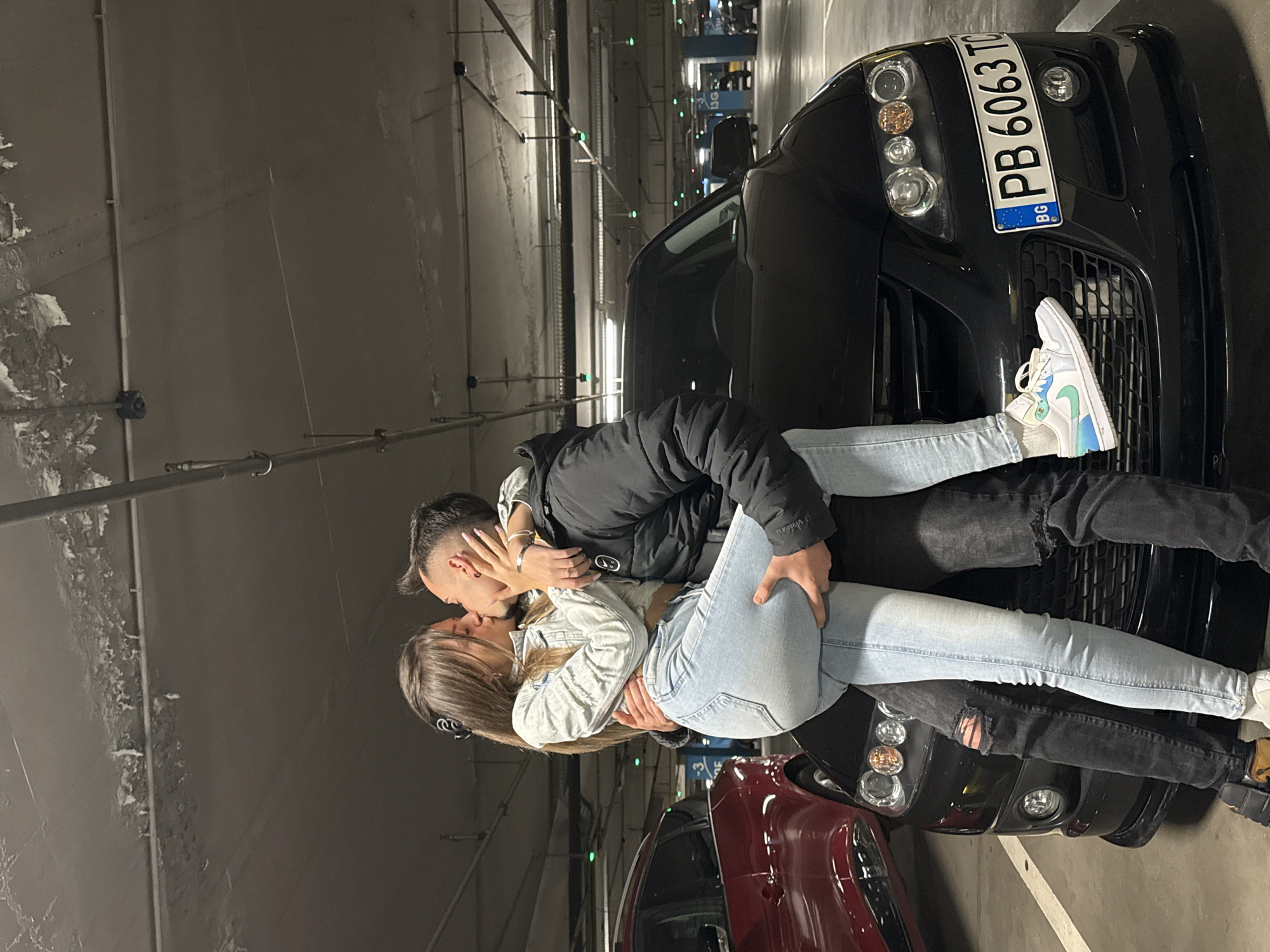 A couple kisses in a parking garage in Sofia, Bulgaria. A woman with long brown hair is wearing a light-colored denim jacket and light blue jeans. The man, whose face is turned away from the viewer, wears a dark jacket and dark jeans. They are standing next to a black SUV with Bulgarian license plate PB6063TC. A red car is parked behind them. The garage has concrete walls and fluorescent lighting.