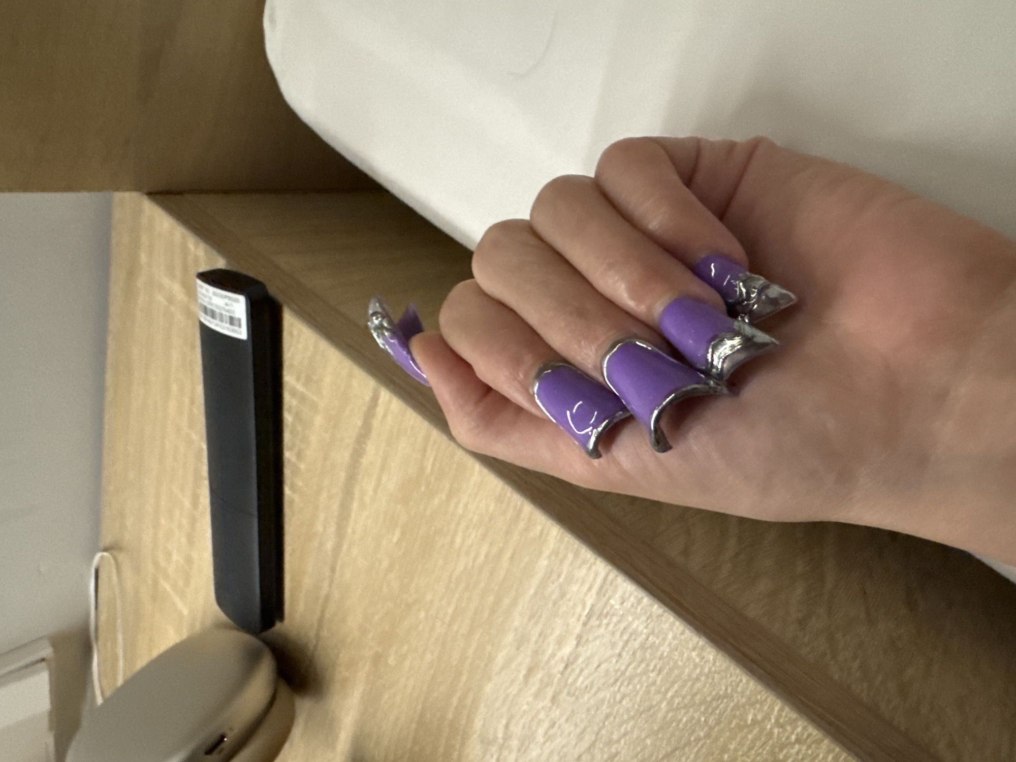The image shows a person's hand with long, purple, pointed acrylic nails adorned with silver accents. The hand rests on a white surface, likely a bed or pillow, next to a wooden bedside table. On the table, a black remote control and a silver electronic device are visible. The background shows a neutral-colored wall. The scene does not provide any specific visual cues to identify its location in Hà Nội, Vietnam.