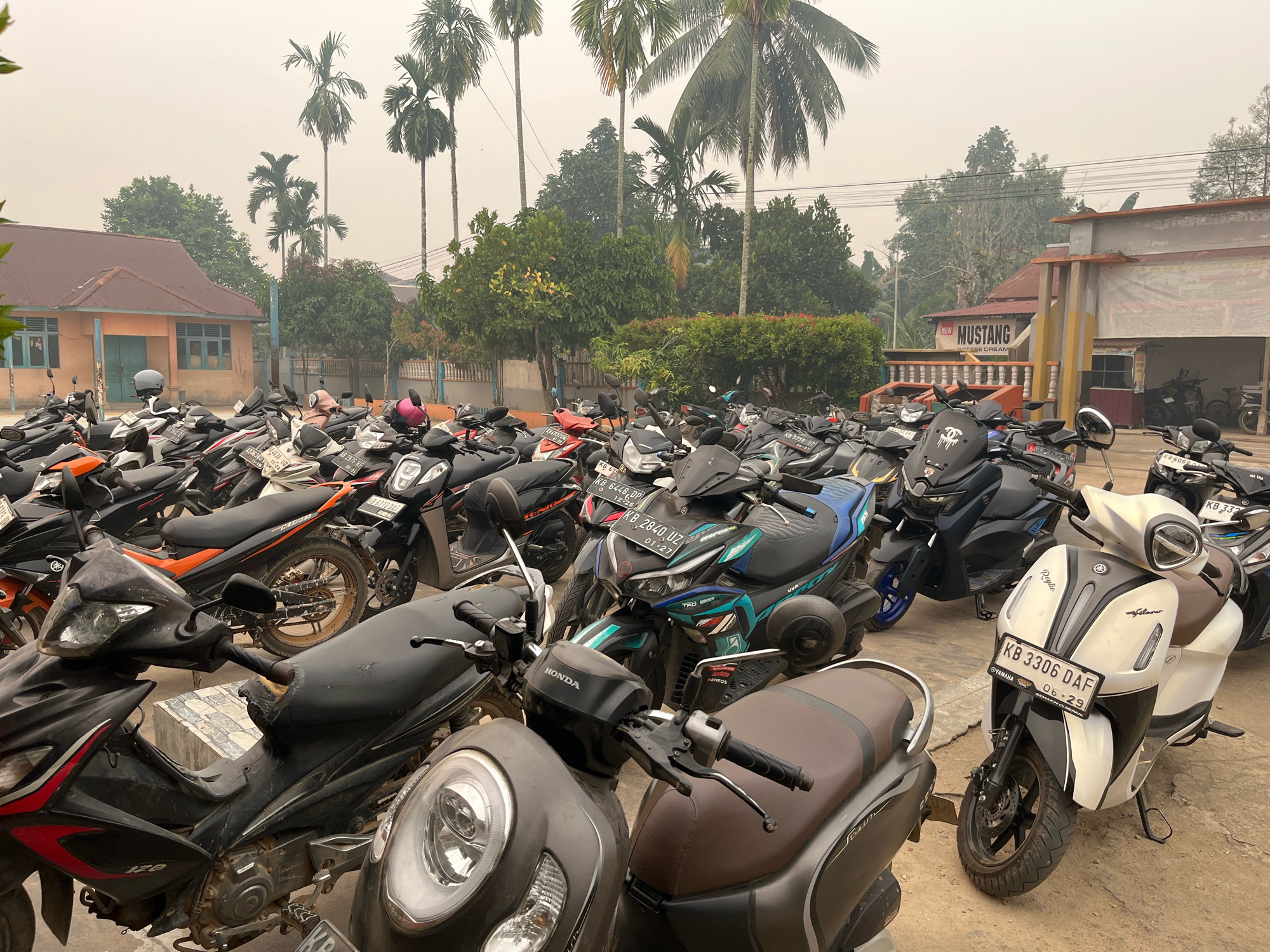 In Meliau Hulu, Indonesia, a parking lot is filled with a large number of parked motorcycles under a hazy, overcast sky. The motorcycles are of various models and colors, predominantly black, white, and orange. Some are sleek and modern, while others appear older. Several are covered with dust, suggesting they have been parked for some time. A few helmets, one pink and one grey, are visible on seats. In the background, a building with an orange facade and a red roof is visible, along with palm trees and other vegetation. The overall scene suggests a public space where people have arrived by motorcycle, possibly for an event or daily activity. The haze in the sky could indicate smoke or pollution.