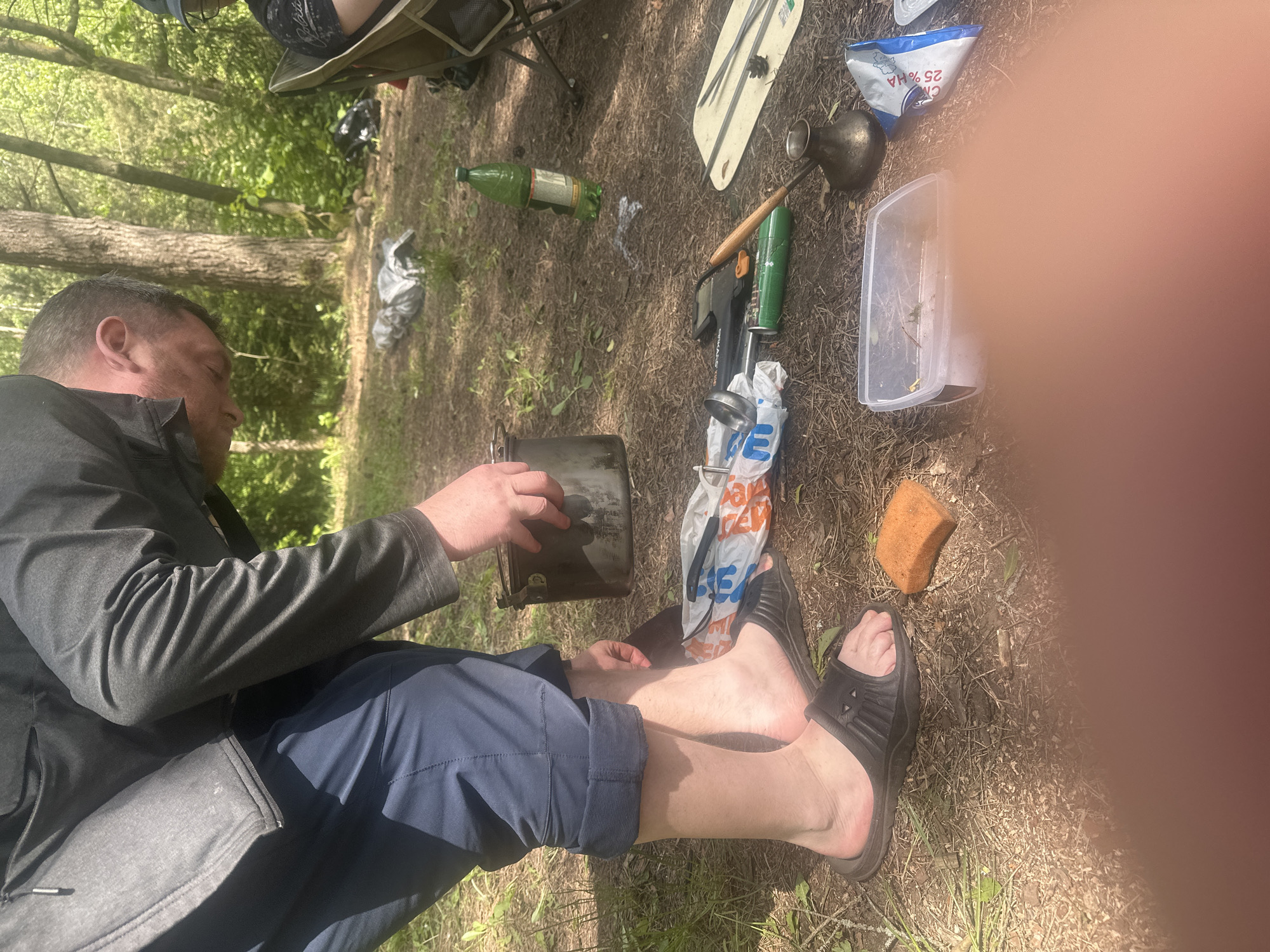 The image, taken outdoors in Папярнянскі сельскі Савет, Belarus, features a man seated on the ground with his legs extended and feet wearing black slide sandals. He is holding a dark-colored camping pot in his hands. The man is dressed in a gray jacket and blue pants. Surrounding him on the ground are various camping-related items, including a green bottle, a white cutting board, a metal pitcher, a green gas canister, a small axe, a plastic bag with a logo, a plastic container with vegetation, and an orange sponge. The ground is covered in dry grass, dirt, and some sparse greenery. A portion of the image is obscured by a blurred orange object.