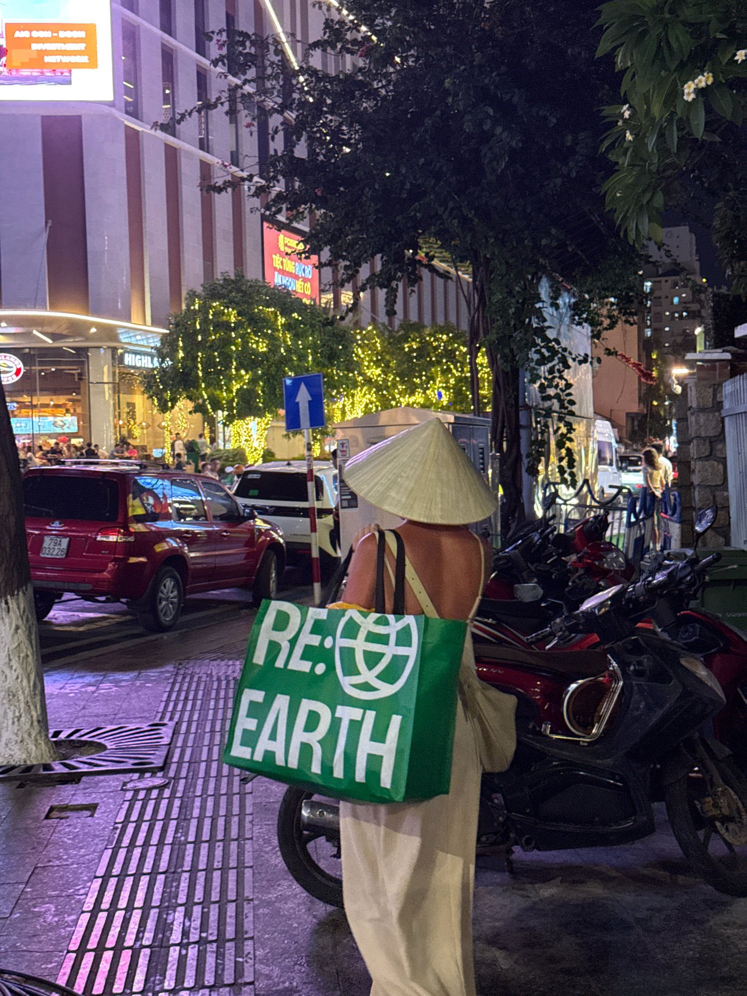 The image depicts a street scene at night in Nha Trang, Vietnam, near the Highlands Coffee shop. A person wearing a conical hat is visible from the back, holding a large green shopping bag that reads "RE: EARTH" in white letters. Behind the person, several red and black motorcycles are parked. In the background, cars are present, including a red SUV with Vietnamese license plates, and a white van. Buildings with illuminated windows and decorative lights are also visible, suggesting a commercial or urban area. The street appears to be paved with textured tiles, and trees with yellow lights strung around them are present, creating a festive atmosphere. The lighting suggests it is nighttime, with artificial lights illuminating the scene.