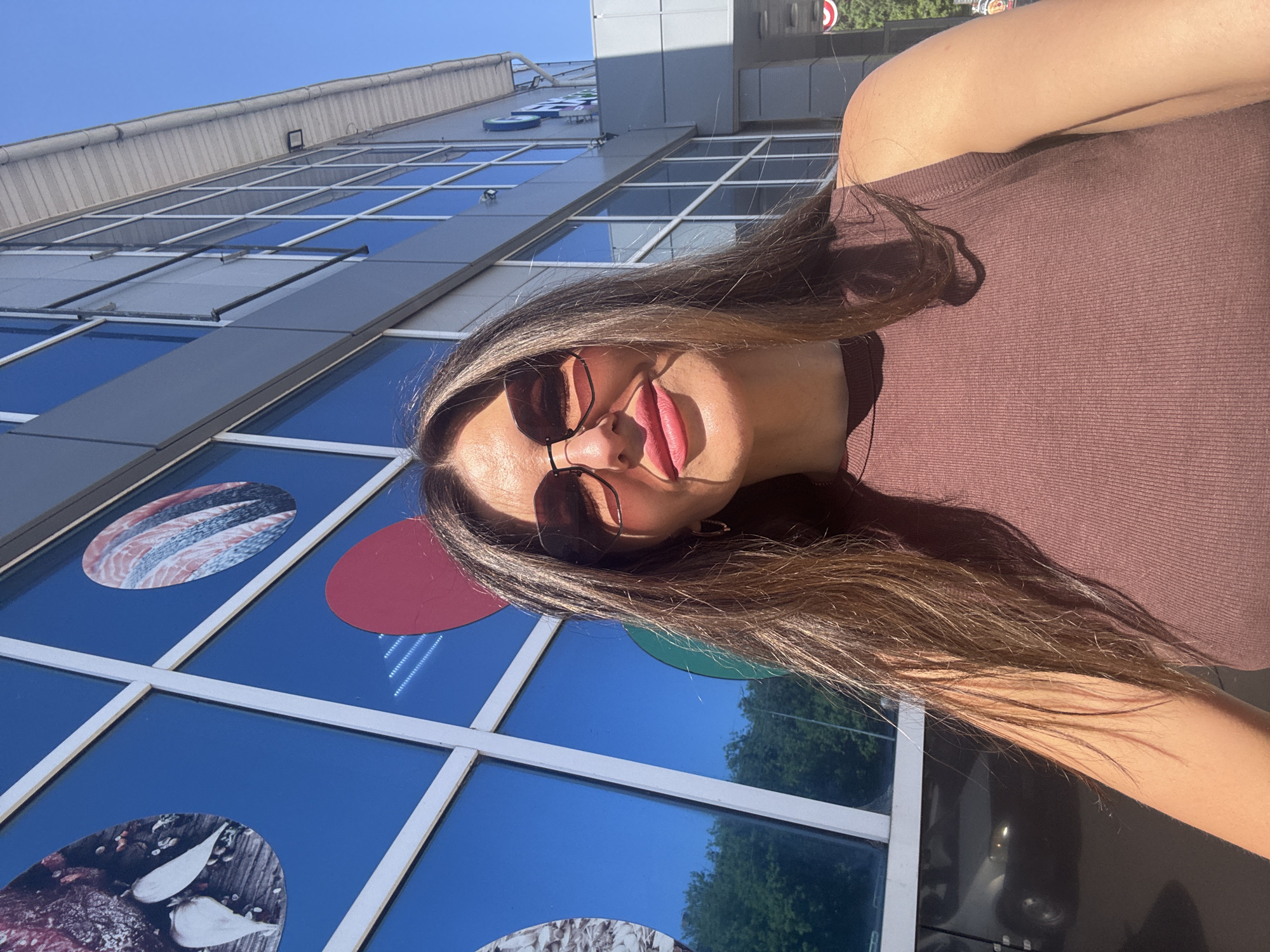 A woman with long brown hair, wearing brown sunglasses and a brown top, stands in front of a building. The building has a facade of large windows decorated with circular stickers featuring food images. The location appears to be in the city of Chaikovsky, Russia, based on the building's design.