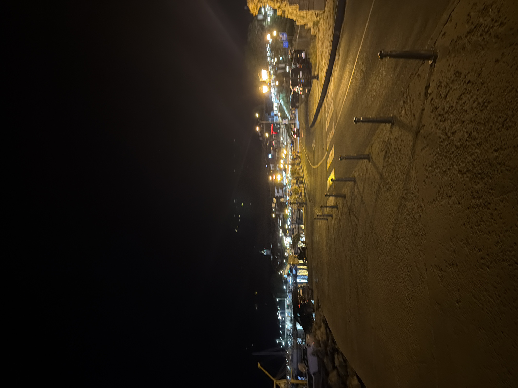 The image depicts a nighttime scene in Nesebar, Bulgaria, likely along a coastal road or promenade. The foreground shows a textured, sandy or gravel path illuminated by streetlights, with a line of bollards separating it from a paved road. The road curves to the right, with streetlights casting a warm glow. Several vehicles, including cars and vans, are visible on the road and parked along its side. In the background, lights from buildings and possibly boats suggest a harbor or town area. The sky is dark, with distant lights hinting at a hillside or farther structures. The overall atmosphere is quiet and serene, characteristic of a seaside town at night.