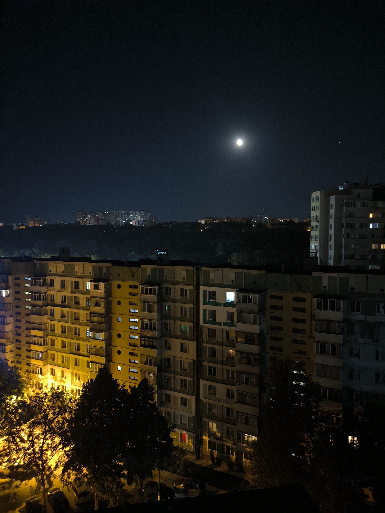 This is a nighttime, high-angle shot of a dense urban environment in Chișinău, Moldova. The scene is dominated by several large, multi-story apartment buildings, typical of Soviet-era architecture, with many windows illuminated from within. In the foreground, the facade of a yellow-toned apartment building is visible, with balconies and windows displaying a warm, artificial light. To its right, a taller, grey apartment building stands, also showing lit windows. In the middle ground, a dark, tree-filled area separates the foreground buildings from a series of apartment buildings in the background, which are also illuminated. The sky above is a deep, dark blue, with a prominent full moon and a few faint stars visible. The overall impression is of a quiet, residential area at night, with the city lights creating a distinct atmosphere.