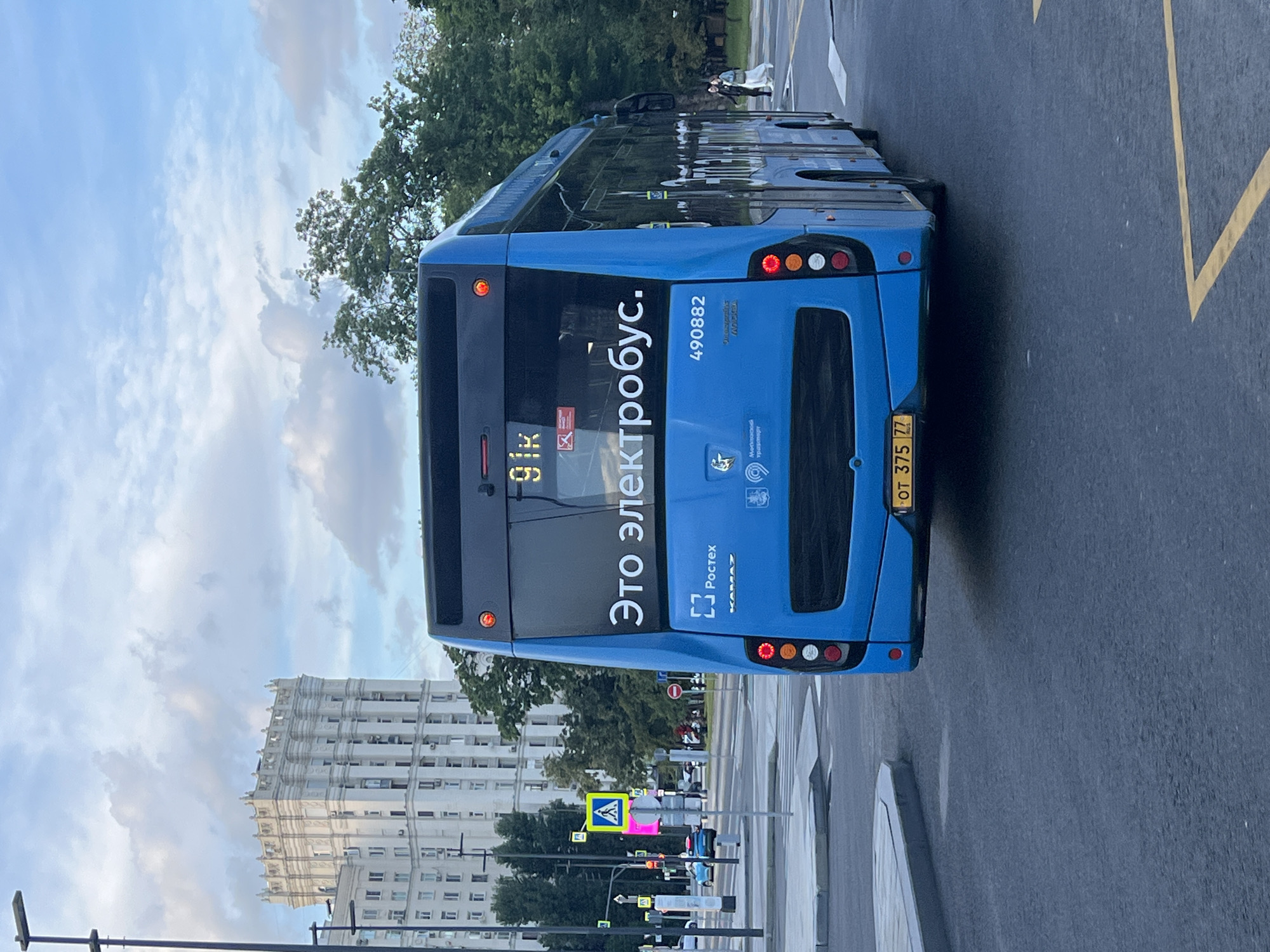 A blue electric bus is parked on a city street. The bus has "Это электробус." (This is an electric bus.) written on the back in white lettering. Below that, the number "490882" is visible. Further down on the left, the word "Ростех" (Rostec) is printed above a logo, and the word "КамАЗ" (Kamaz) is printed below it. On the right side of the bus, a license plate reads "ОТ 375 77".
The bus is oriented vertically in the image, with the rear facing the camera. It is daytime, and the sky is partly cloudy with patches of blue. Trees and a tall, ornate white building are visible in the background. A pedestrian crossing with yellow markings is painted on the road in front of the bus, and a yellow pedestrian crossing sign is displayed. Several traffic lights and street signs are also present. The overall scene appears to be a typical urban environment.