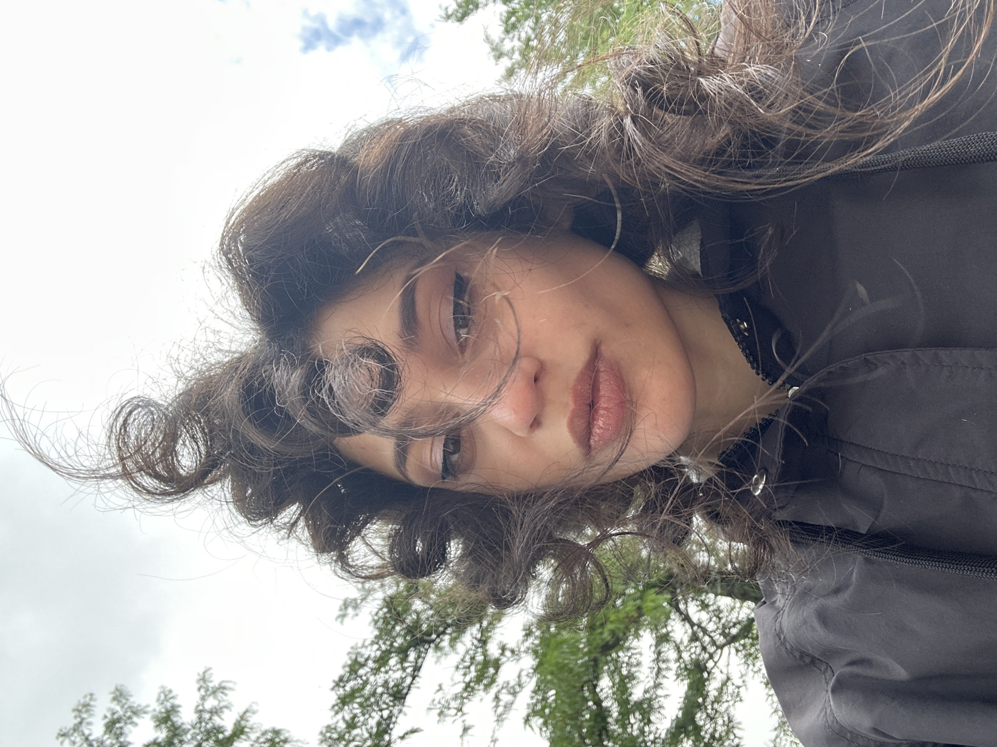 The image is a selfie taken outdoors with a cloudy sky and trees in the background. The subject is a young woman with dark, curly hair that is windblown. She is wearing a black jacket. Her face is partially obscured by her hair. The photo is taken from a low angle, looking up at the woman. The location in the city of Sofia, Bulgaria cannot be determined from the image.