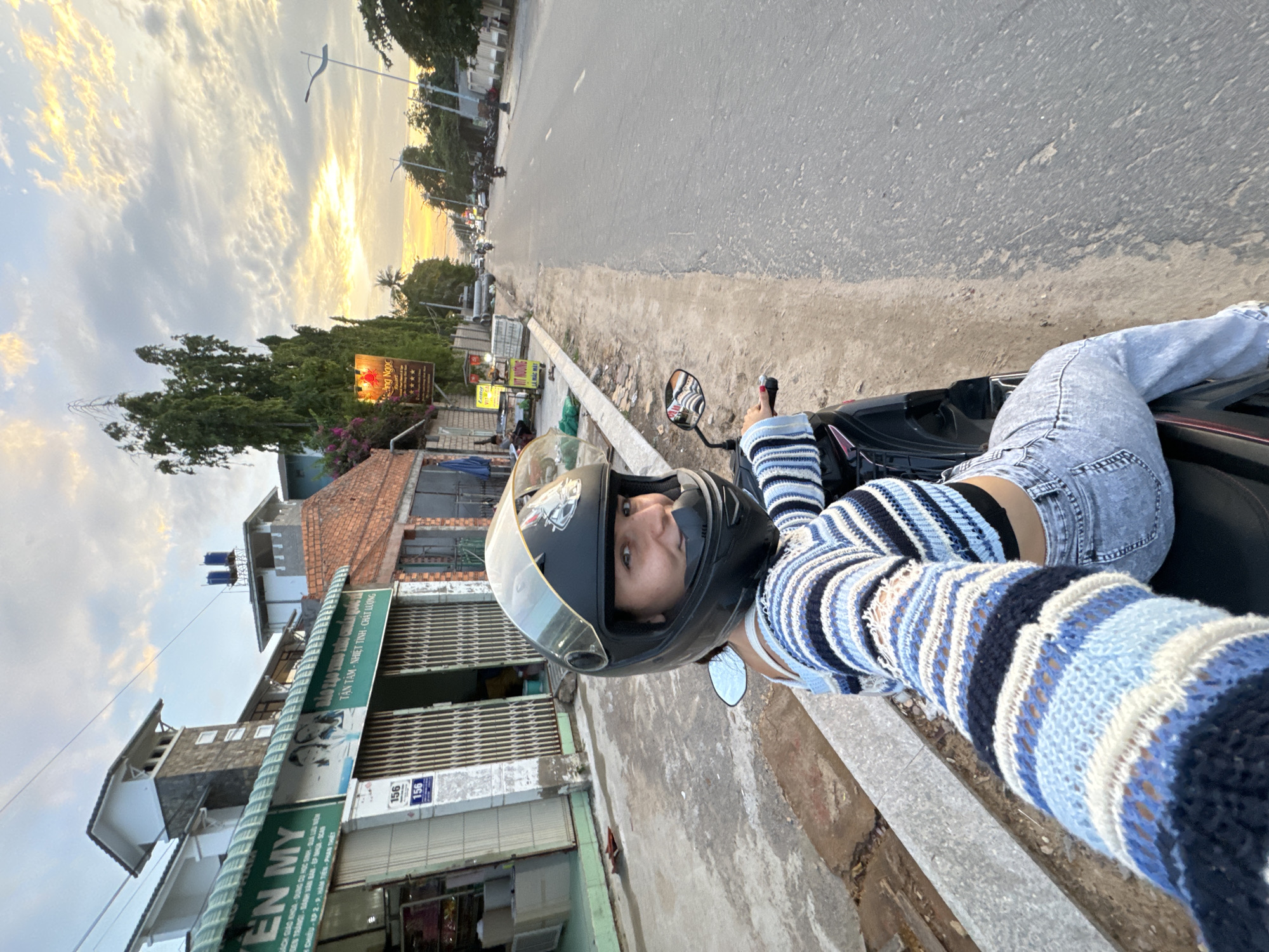 A young woman wearing a black helmet and a blue and white striped sweater is riding a motorcycle. She is wearing distressed denim jeans and is looking at the camera. The motorcycle is black and parked on the side of a road. The scene is set in Mũi Né, Vietnam, during what appears to be sunset. The sky is filled with dramatic clouds, tinged with warm yellow and orange hues.
To the left of the road, there are buildings with brick roofs and signs with Vietnamese text. One prominent green sign reads "TÂN TÂM - NHIỆT TÌNH - CHẤT LƯỢNG" which translates to "Dedicated - Enthusiastic - Quality". Below this, another sign partially visible reads "EN MY" and other Vietnamese text including "BÌNH VĂN BẢN". There are also numbers "156" visible on some of the buildings. Trees and other vegetation line the side of the road, with some flowering bushes adding color to the scene. The road itself appears to be a main thoroughfare with traffic visible in the distance. The overall mood is casual and adventurous, suggesting a scenic ride in the late afternoon.