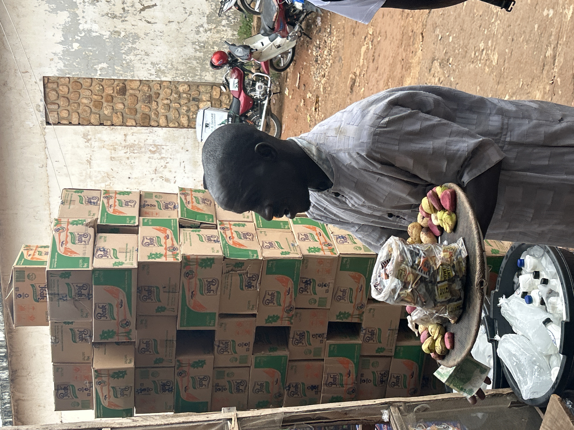 Here's a detailed description of the image:
A man stands next to a large stack of boxes labeled "Peak" (likely boxes of powdered milk) in what appears to be a small shop or market. The boxes are stacked in a pyramid shape. The man is holding a shallow, round tray with various items for sale, including small bags of candy, peanuts, and what seems to be bottled drinks in a black bucket. A few Naira bills are visible on the tray.
Behind the man, in the background, is a wall with a textured stone or brick section. A motorcycle is also visible in the background.
Based on the context provided in the prompt, it's reasonable to infer that this scene is taking place in Jos, Nigeria. The presence of Naira currency, the "Peak" brand (common in West Africa), and the general setting all support this location.