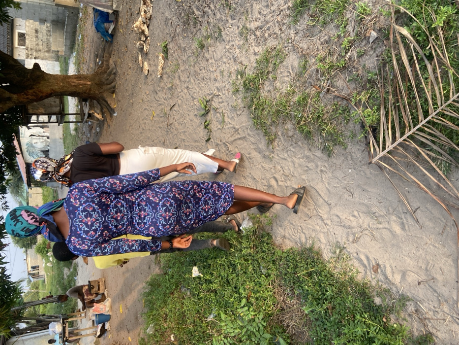 Four individuals stand on a sandy, grassy area. Two women are prominently featured: one in a navy blue floral dress and head wrap, and another in a black shirt and white pants, also with a head wrap. Two other people in a yellow shirt and light-colored shirt are partially visible. A large tree with exposed roots stands nearby, with a building faintly visible in the background. Debris, including husks, litters the sandy ground. A dried palm frond rests on the sand.