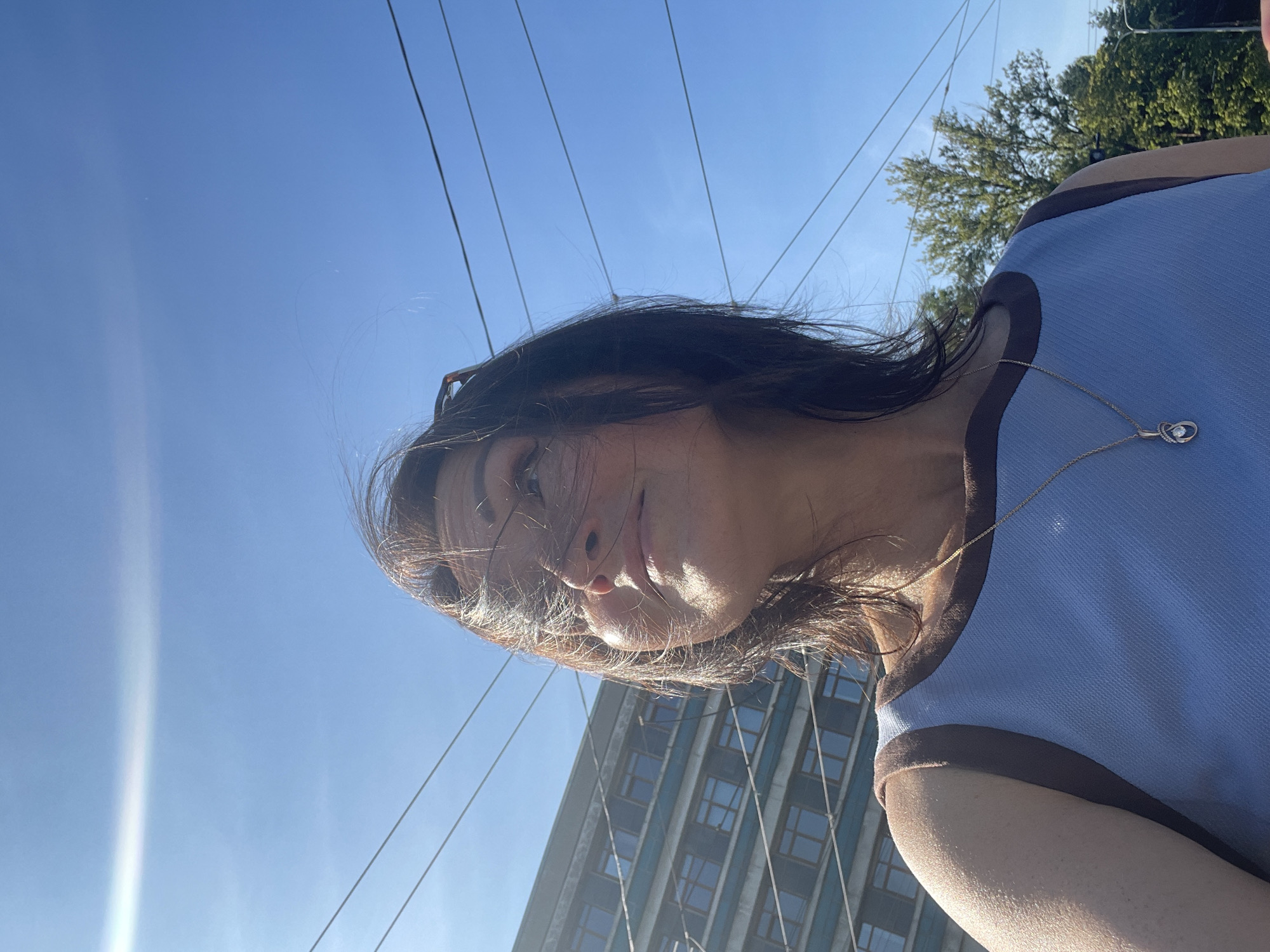 A woman is captured from a low angle outdoors, looking up towards a clear blue sky with some wispy clouds. Her hair is blowing in the wind. She is wearing a light blue tank top with brown trim and a gold necklace with a pendant. In the background, a multi-story building with many windows is visible, along with overhead wires, suggesting an urban environment. The lighting indicates it is daytime, likely sunny. The scene is consistent with an outdoor setting in Saint Petersburg, Russia, possibly near a street with overhead tram or trolleybus lines and apartment buildings.