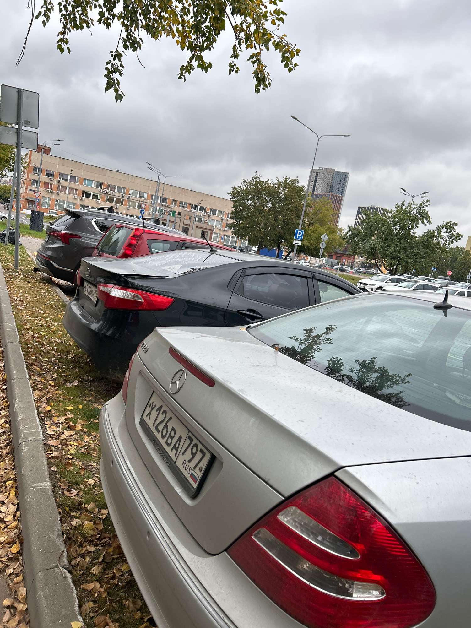 The image captures a street-level view of a parking lot with several cars parked parallel to a curb on an overcast day. The foreground is dominated by the rear of a light-colored sedan, likely a Mercedes-Benz based on the emblem visible on the trunk. Its license plate, with Russian characters and numbers "X126BA 797", is clearly legible. To the left of this vehicle, fallen autumn leaves are scattered on the grass and the edge of the sidewalk. Further back, a dark-colored sedan is parked, and behind it, a red SUV and a dark gray SUV are also visible. In the background, a multi-story building with a facade of light-colored panels and numerous windows stretches across the frame. Further in the distance, modern high-rise buildings are visible against the cloudy sky. Streetlights and traffic signs, including a blue parking sign, are also present in the scene. The overall impression is an urban parking area in Moscow, Russia, during the autumn season.