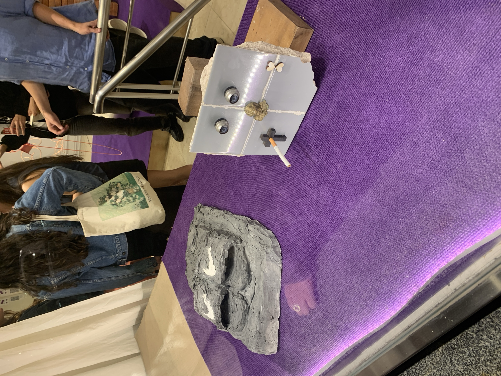 The image displays an indoor scene, likely an art exhibition or gallery, characterized by several individuals observing displayed art pieces. The primary display surface is covered in a vibrant, textured purple fabric or carpet.
Several people are visible in the foreground and mid-ground. One person, seen from behind, has long, wavy dark hair and is wearing a denim jacket over a black top. They carry a white canvas tote bag with the text "VINCENT VAN GOGH" and "ARTOLOGY" clearly visible. Another individual, dressed in a light blue collared shirt, is partially seen near a silver metal railing. Further back, more partial figures of people are discernible, suggesting a populated event.
Two distinct art objects are prominently displayed on the purple surface. The central piece consists of a fractured, light grey slab, possibly concrete or tile, seemingly broken into four sections. This piece is adorned with diverse elements: two small, silver-toned, dome-shaped components, a golden, ornate embellishment that resembles a cherub's head or decorative seal, and a black cross-like object from which a cigarette with a filter protrudes. A small, light-colored butterfly-shaped adornment is also affixed to the slab. This assemblage rests upon two unrefined wooden blocks. Adjacent to this, there is an irregularly shaped, dark grey, textured sculpture, resembling a molded rock or organic form, featuring multiple deep indentations and some white painted accents. A bare toe, wearing a ring, is incidentally visible on the purple fabric near this sculpture.
The setting implies an active cultural event, potentially an art show in Buenos Aires, Argentina, given the context. The lighting suggests an indoor environment, likely under artificial illumination. The objects' arrangement and the observers' presence create an atmosphere of artistic appreciation or contemplation.