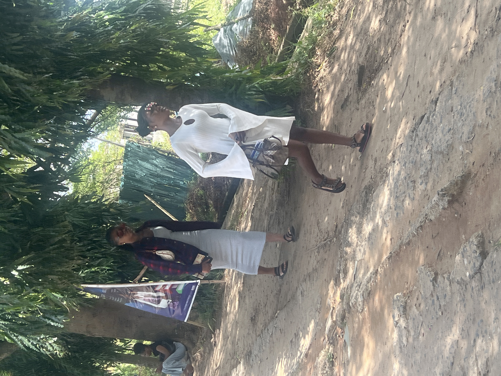 The scene depicts an outdoor location in Ijanikin, Nigeria, characterized by a dirt road lined with trees and foliage. There are two women in the photo. One wears a white dress with flared sleeves and sandals, holding a patterned bag. The other wears a grey dress with a plaid jacket and sandals, holding what appears to be a mobile phone. A banner or sign is mounted near a tree. In the background, there is a structure covered in blue tarpaulin. Another person is visible near the tree at the bottom of the frame.