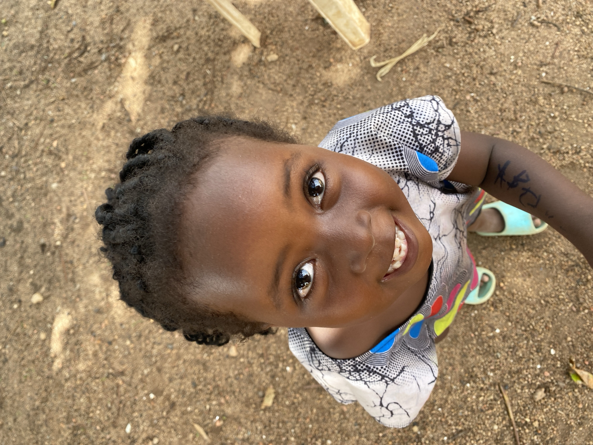 A high-angle, close-up photograph captures a young girl from Kuru Dungus, Nigeria, looking directly up at the camera with a wide, joyful smile. Her dark, expressive eyes are bright, and her face is illuminated, highlighting her cheerful demeanor and prominent front teeth. Her hair appears to be styled in short, dark braids or twists.
She is wearing a patterned top with a light background featuring black geometric shapes, dots, and abstract lines, accented with splashes of blue, yellow, and red. On her left forearm, a dark, crudely drawn symbol resembling "APC" or similar letters is visible. Her feet, clad in light blue and green open-toe sandals, are partially visible at the bottom right.
The setting is outdoors, with the background consisting of sandy or dirt ground speckled with small pebbles, dried leaves, and scattered light-colored debris, suggesting a natural or unpaved environment. The lighting indicates it is daytime. The overall mood of the image is one of innocence, happiness, and direct engagement with the viewer.