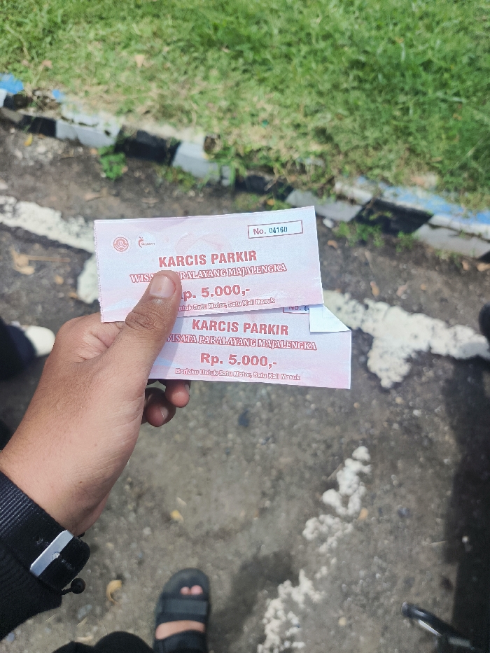 A person is holding a parking ticket from the "Wisata Paralayang Majalengka" in Majalengka, Indonesia (latitude: -6.846, longitude: 108.204). The ticket is pink and white, with black text. The text reads, "Karcis Parkir Wisata Paralayang Majalengka Rp. 5.000,- Berlaku Untuk Satu Motor Satu Kali Masuk." The person is wearing a black shirt with a black watch. The scene is taken from a downward angle and shows the person's hand holding the ticket and their left foot wearing a brown sandal. The ground is a mix of dirt and concrete. The background is blurred and shows green vegetation.