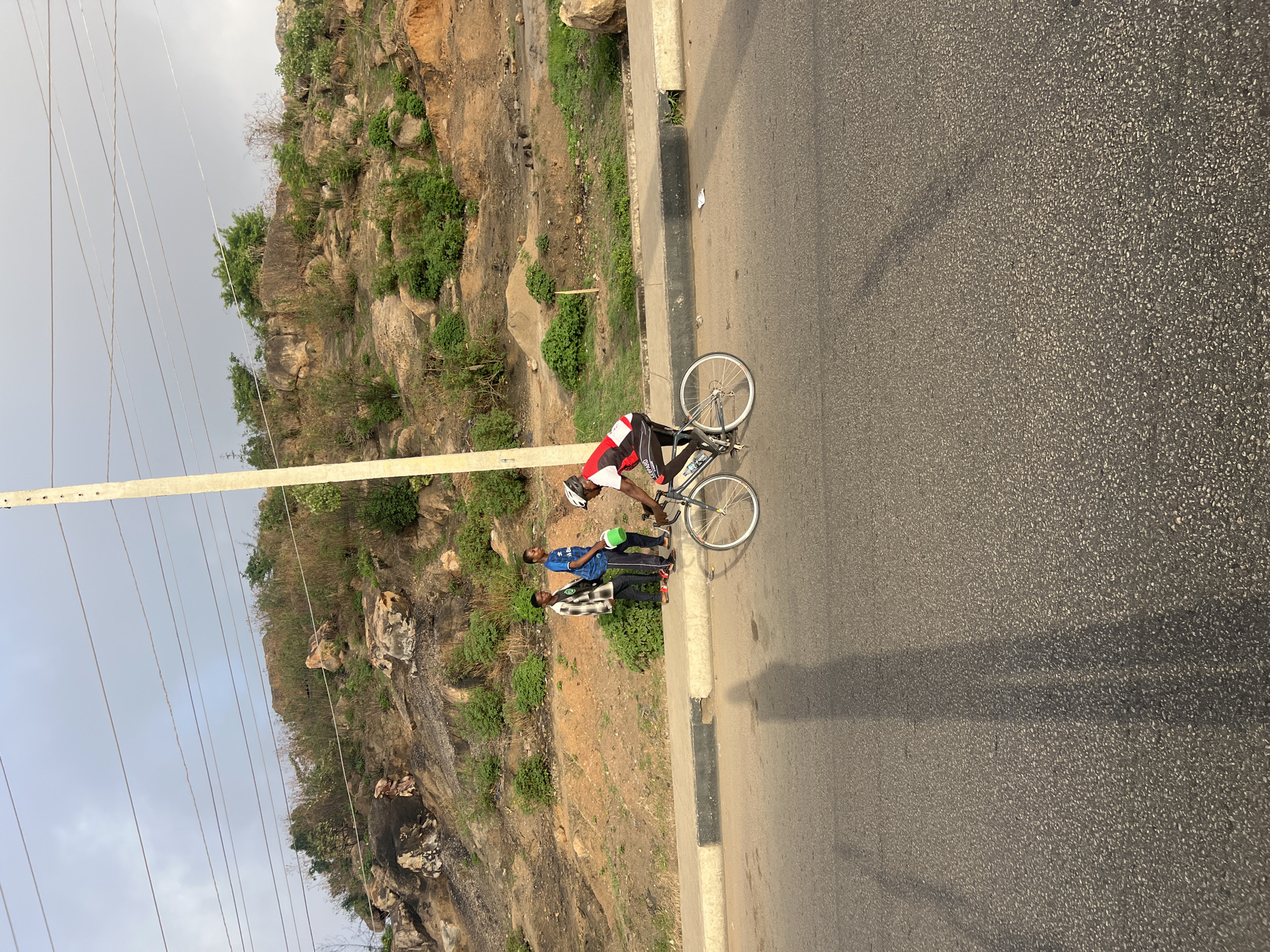 A cyclist with a white helmet and red-and-white athletic gear is standing with his bicycle on the shoulder of a paved road in Gura Top, Nigeria. Two men stand nearby. A rocky hillside with scrub vegetation rises sharply to the left. A utility pole with power lines runs along the hillside. The sky is overcast.