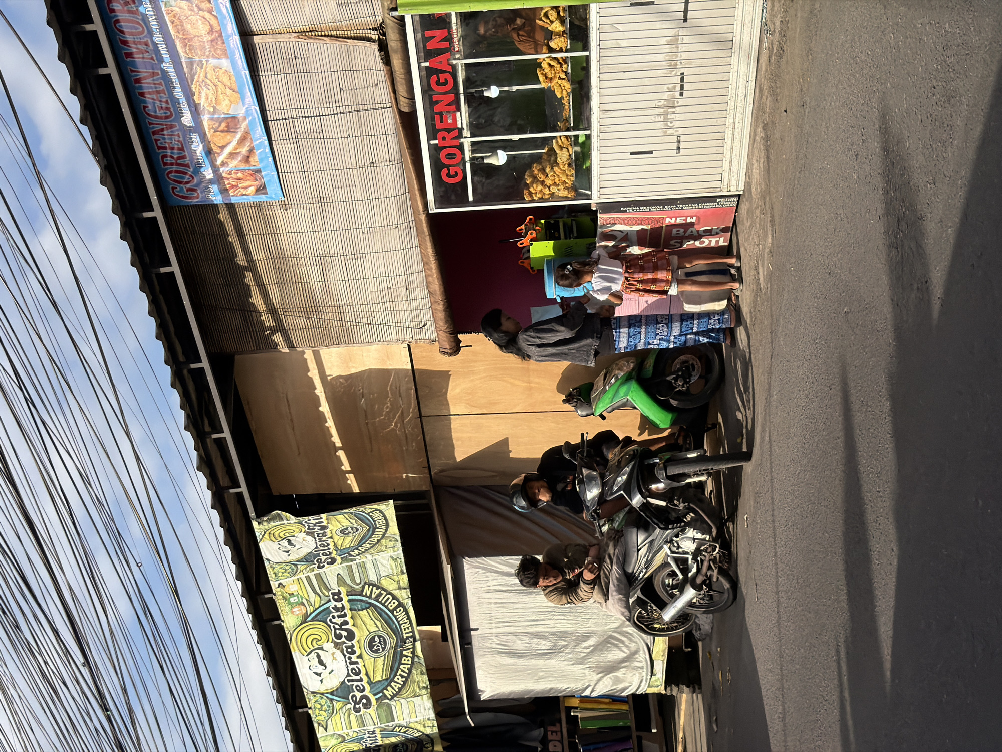 In an outdoor street scene under a bright sky cluttered with overhead wires, two individuals sit on a motorcycle parked on the asphalt. To the right of the motorcycle, a food stall with a glass display case showcases fried foods. A woman stands by the stall, attending to a young girl dressed in a patterned skirt and white top. A green motorcycle is parked behind the food stall. Banners with text and illustrations are hung above the stall and to the left. The overall impression is a vibrant street-side food vendor operation.