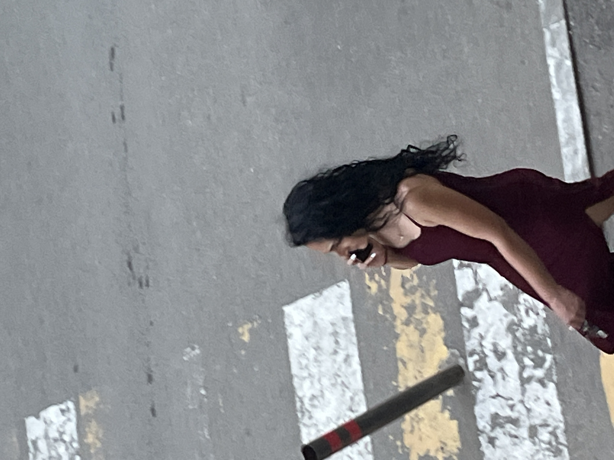 A woman with long, dark, wavy hair, wearing a dark red dress, is seen from the side. She is holding a mobile phone to her ear and appears to be speaking. The background is an asphalt surface with white and yellow markings, suggestive of a pedestrian crossing or road marking. A dark cylindrical object, possibly a pole or barrier with red stripes, is visible in the foreground, angled diagonally across the frame. The lighting suggests it is daytime, and the overall scene appears to be outdoors in an urban environment. No text is visible in the image.