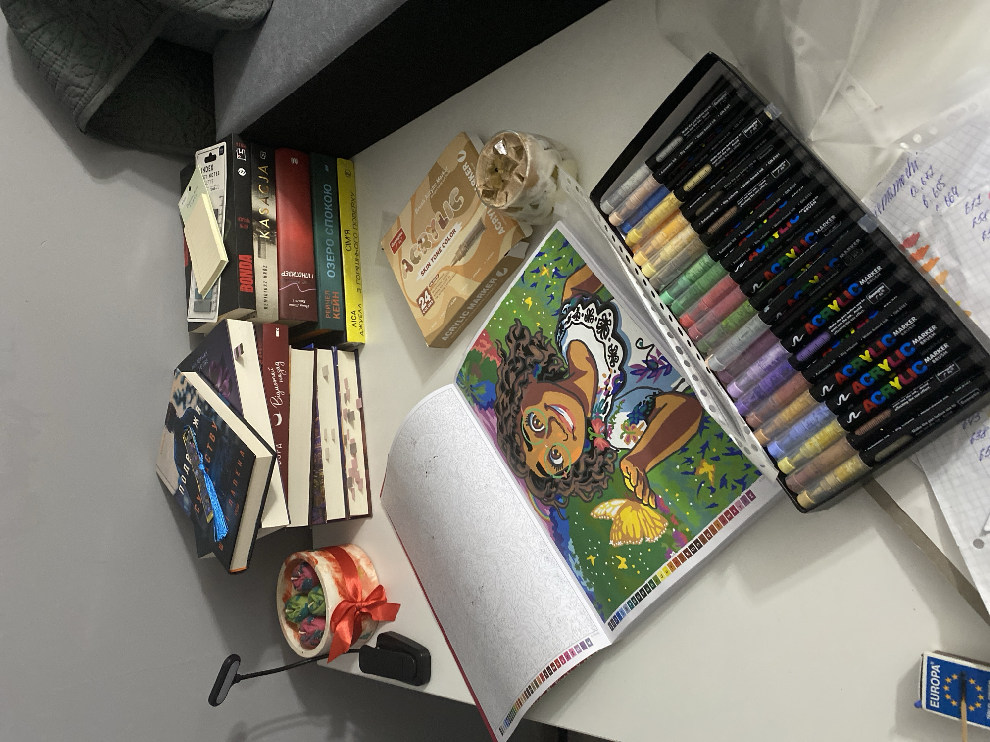 A stack of books sits on a desk next to a small decorative pot with green and pink candles tied with a red ribbon. To the right of the books, a box of "ACRYLIC MARKER" pens and a glass container with small decorative objects are visible. A large open book, a paint-by-numbers coloring book, lies on the desk, displaying a colorful illustration of a character and a butterfly, with an uncolored page next to it. A box of "EUROPA" matches is also on the desk. The scene is indoors, with a grey wall in the background and a portion of a gray sofa visible on the right. The overall impression is of a creative workspace. The text on the books and pens are primarily in Polish.